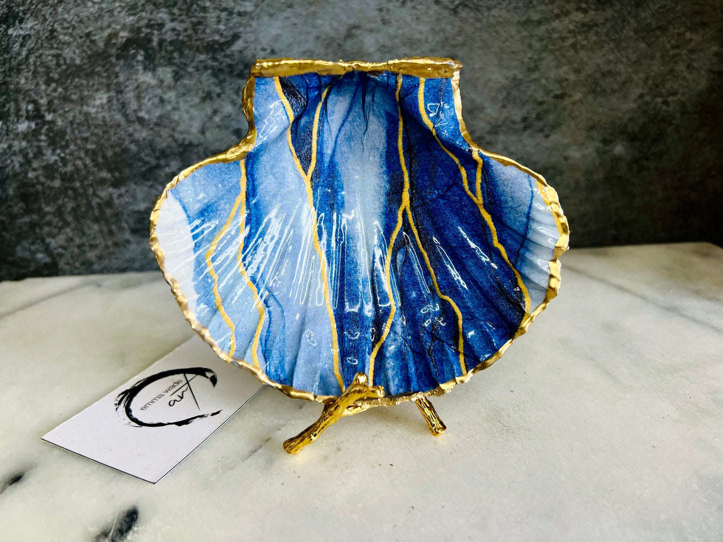 Hand painted blue waves scallop shell trinket dish with gold leaf accents and glossy resin finish, shown with optional gold driftwood display stand. Coastal jewellery and ring dish crafted from a natural Dorset scallop shell, featuring flowing blue wave artwork. Decorative shell bowl photographed on a light background, showing the natural shell shape and gilded edges, ideal for holding rings, earrings, trinkets or small treasures.