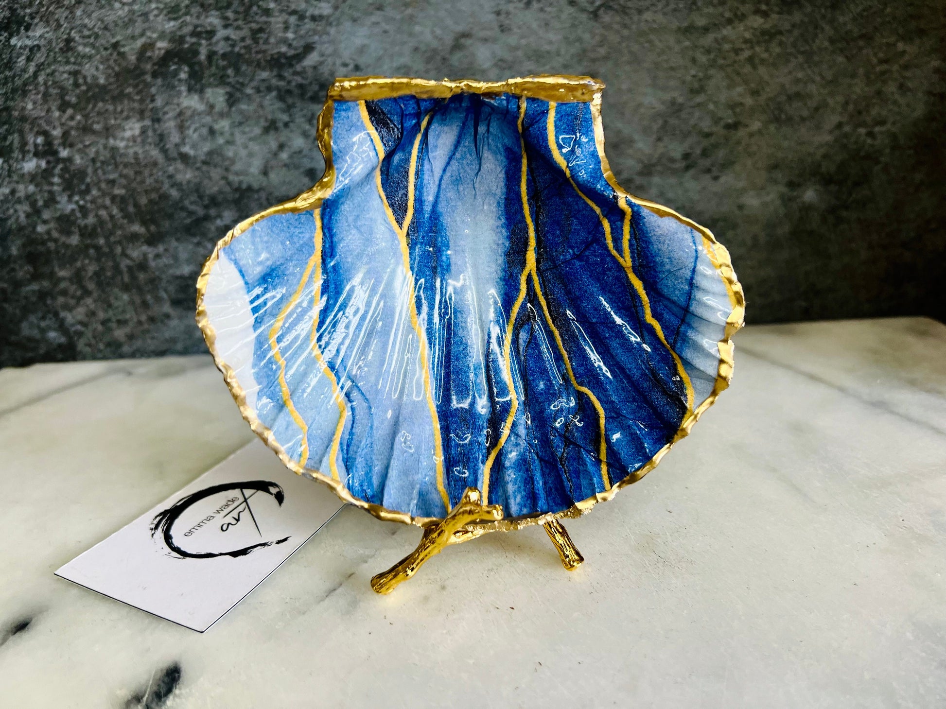Hand painted blue waves scallop shell trinket dish with gold leaf accents and glossy resin finish, shown with optional gold driftwood display stand. Coastal jewellery and ring dish crafted from a natural Dorset scallop shell, featuring flowing blue wave artwork. Decorative shell bowl photographed on a light background, showing the natural shell shape and gilded edges, ideal for holding rings, earrings, trinkets or small treasures.