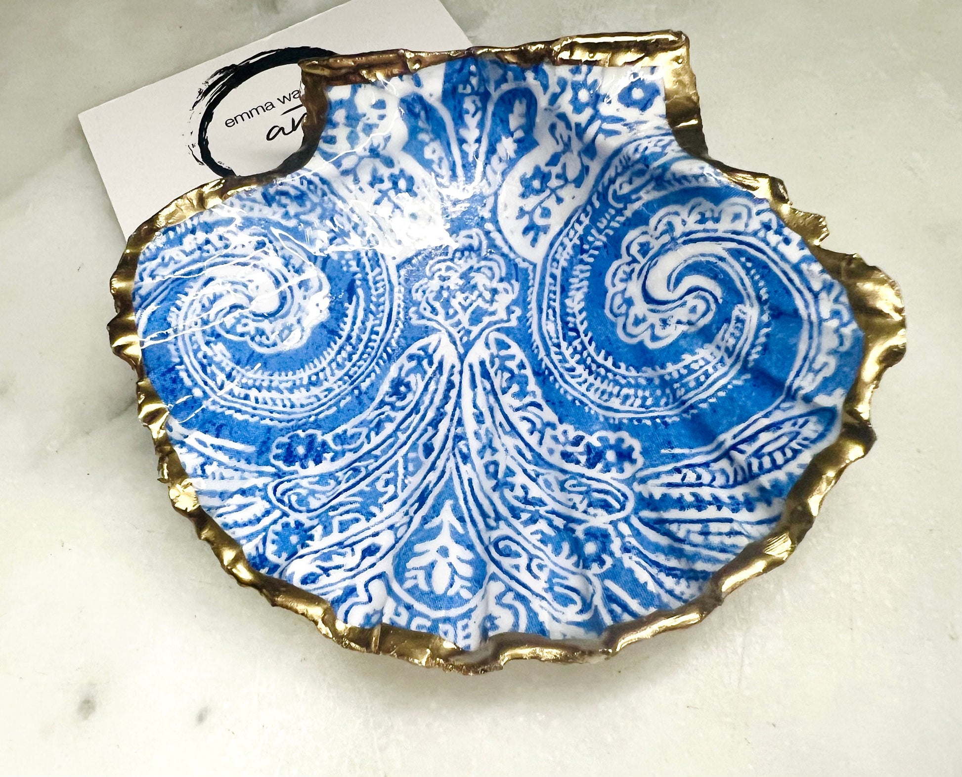 Blue and white paisley scallop shell ring catcher with hand applied gold leaf edging and glossy resin finish. Handmade Dorset shell jewellery tray styled with pearls on a marble background. Coastal vanity decor and luxury beach house accessory gift.