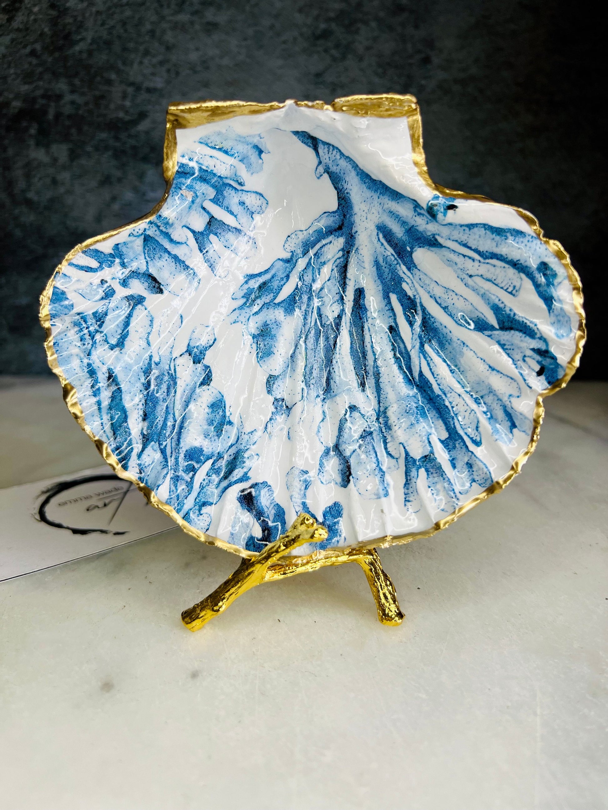 Blue seaweed scallop shell ring catcher with hand applied gold leaf edging and glossy resin finish. Handmade Dorset shell jewellery tray with ocean inspired design styled on marble background. Coastal vanity decor and beach house accessory with optional gold driftwood stand.