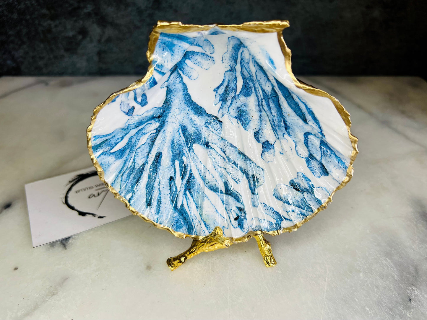 Blue seaweed scallop shell ring catcher with hand applied gold leaf edging and glossy resin finish. Handmade Dorset shell jewellery tray with ocean inspired design styled on marble background. Coastal vanity decor and beach house accessory with optional gold driftwood stand.