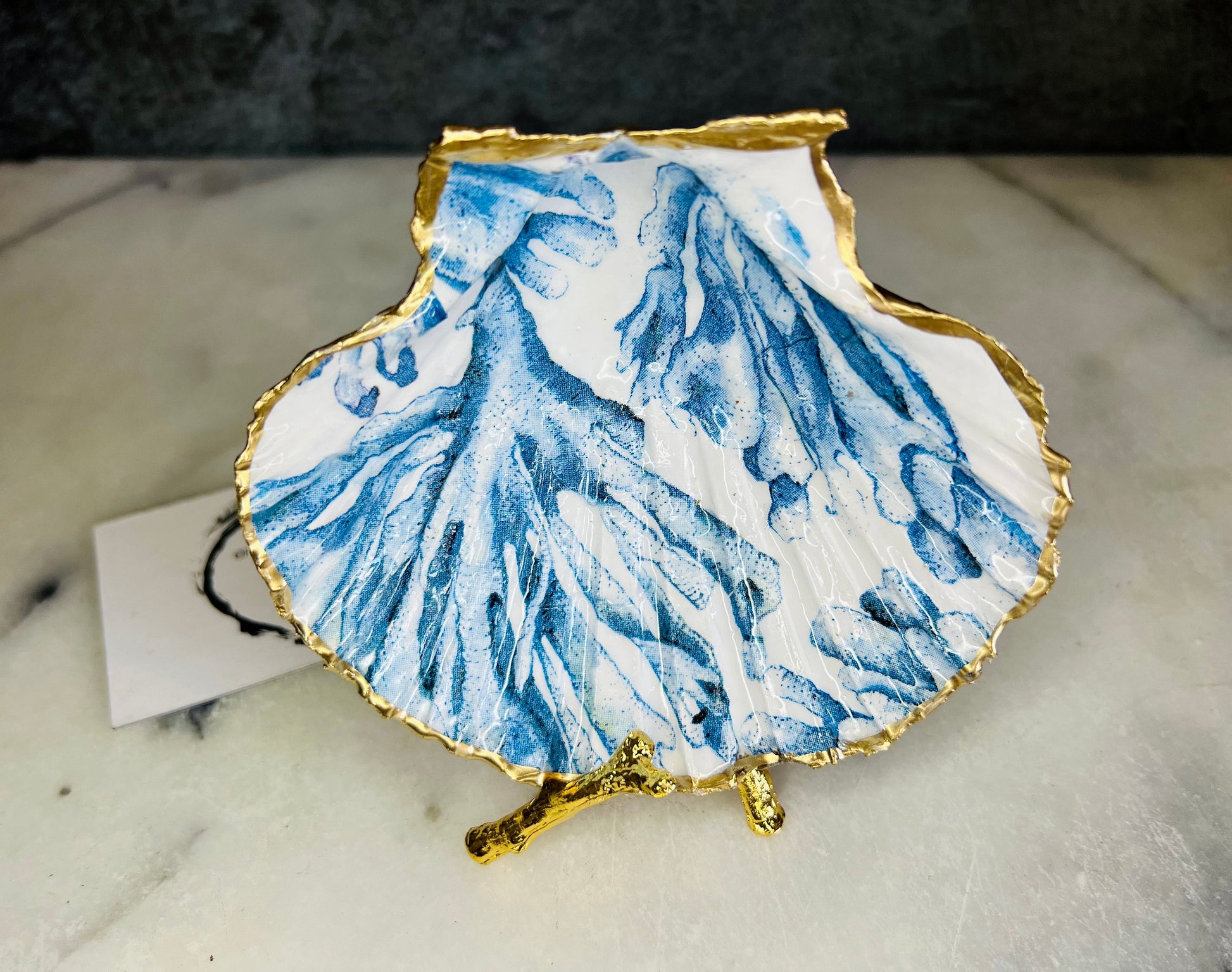 Blue seaweed scallop shell ring catcher with hand applied gold leaf edging and glossy resin finish. Handmade Dorset shell jewellery tray with ocean inspired design styled on marble background. Coastal vanity decor and beach house accessory with optional gold driftwood stand.