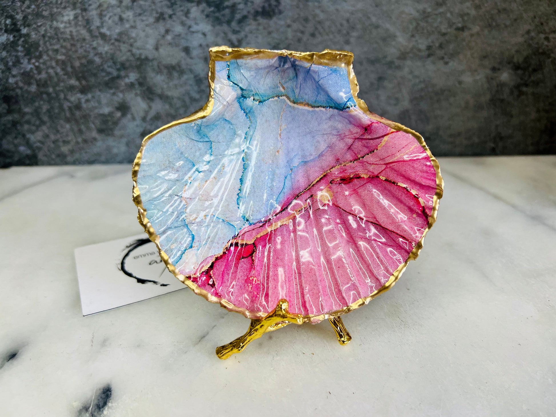 A handmade scallop shell trinket dish painted in soft pink and pale blue tones in a flowing, watercolor-style pattern, finished with a gold leaf edge and sealed with a glossy clear resin. The natural shell shape is visible, with the ridged texture of the scallop shell showing through the design. The dish is shown on a light surface and works as a small decorative tray suitable for holding rings or jewellery in a bedroom or bathroom.