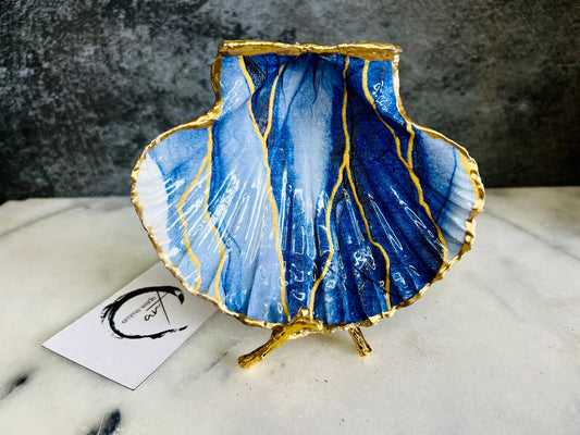 Hand painted blue waves scallop shell trinket dish with gold leaf accents and glossy resin finish, shown with optional gold driftwood display stand. Coastal jewellery and ring dish crafted from a natural Dorset scallop shell, featuring flowing blue wave artwork. Decorative shell bowl photographed on a light background, showing the natural shell shape and gilded edges, ideal for holding rings, earrings, trinkets or small treasures.