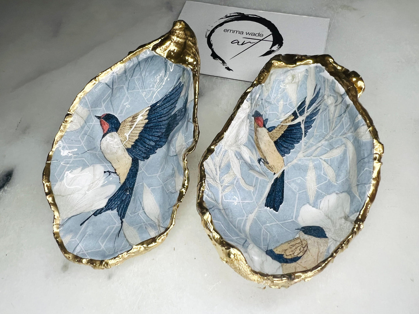 Two natural oyster shell salt and pepper dishes decorated with a blue bird decoupage design, finished with hand-gilded gold leaf edges and sealed with glossy resin. Shown with small gold spoons, these handmade coastal shell pinch pots are crafted from Dorset shells and styled as an elegant table setting accessory or unique gift.