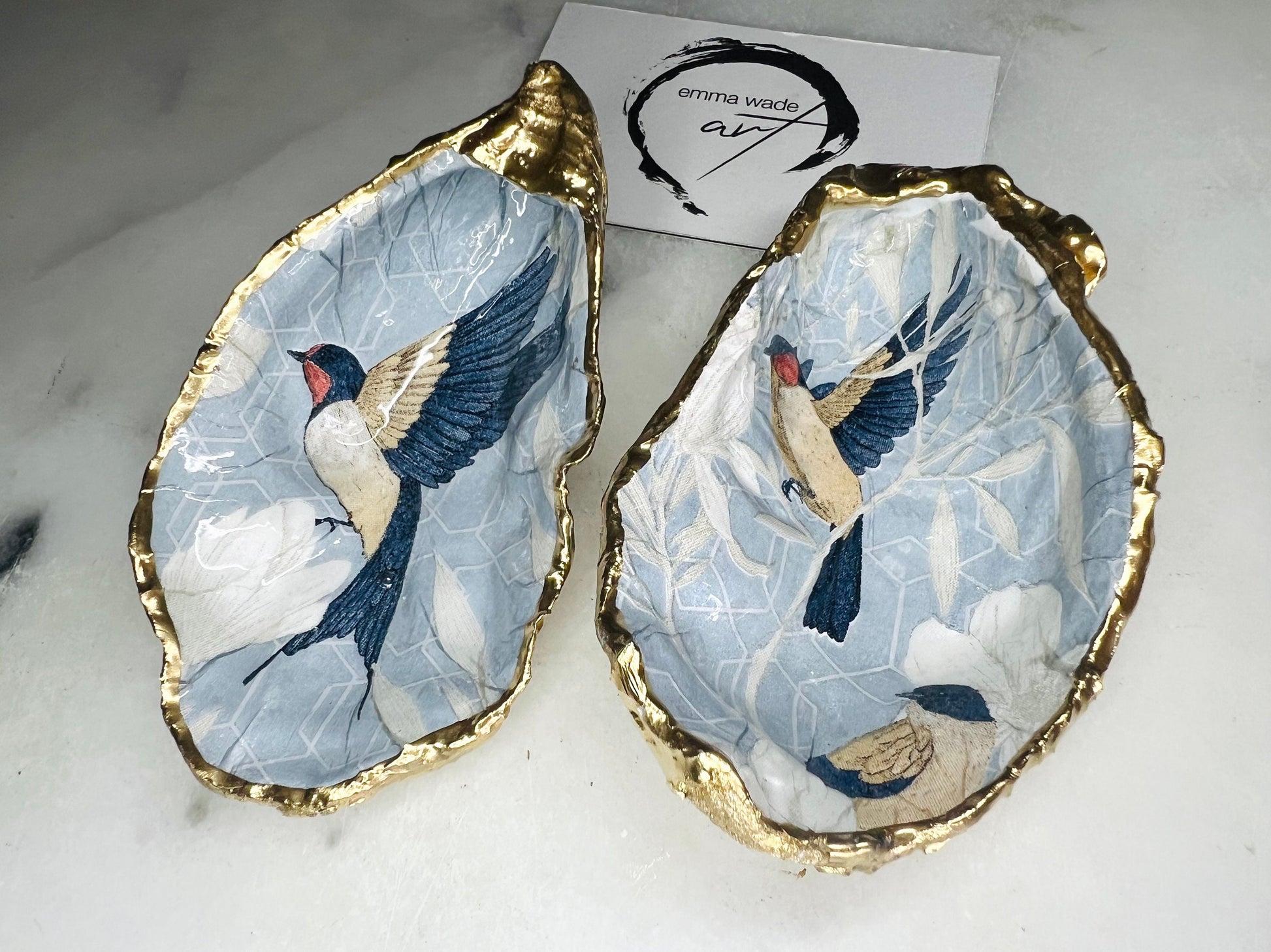 Two natural oyster shell salt and pepper dishes decorated with a blue bird decoupage design, finished with hand-gilded gold leaf edges and sealed with glossy resin. Shown with small gold spoons, these handmade coastal shell pinch pots are crafted from Dorset shells and styled as an elegant table setting accessory or unique gift.