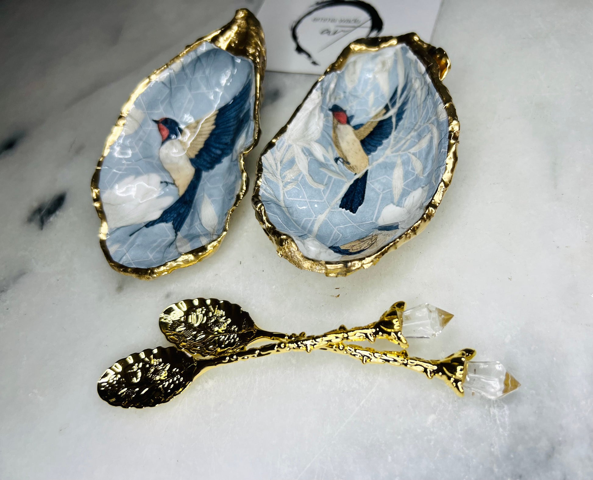 Two natural oyster shell salt and pepper dishes decorated with a blue bird decoupage design, finished with hand-gilded gold leaf edges and sealed with glossy resin. Shown with small gold spoons, these handmade coastal shell pinch pots are crafted from Dorset shells and styled as an elegant table setting accessory or unique gift.