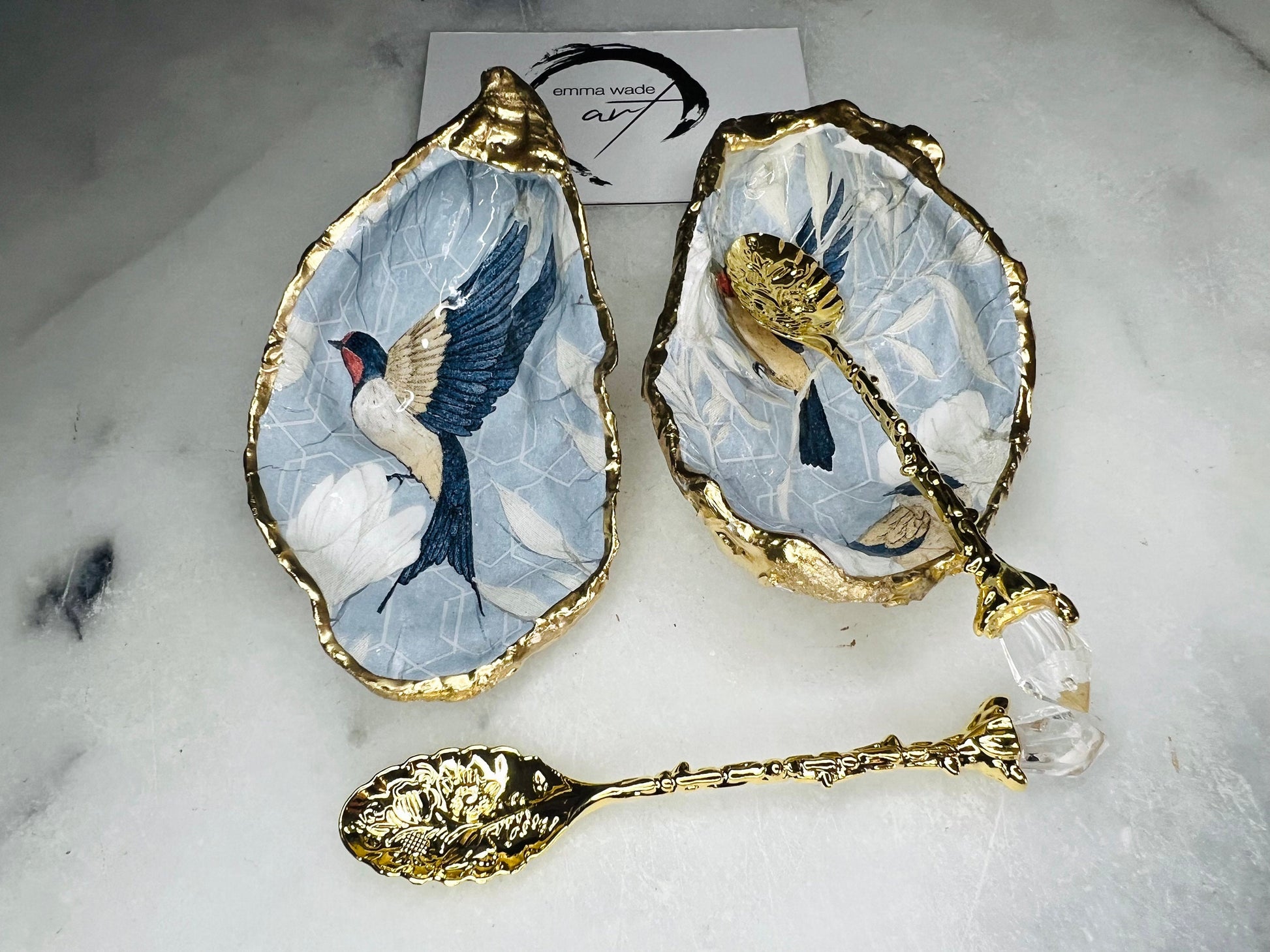 Two natural oyster shell salt and pepper dishes decorated with a blue bird decoupage design, finished with hand-gilded gold leaf edges and sealed with glossy resin. Shown with small gold spoons, these handmade coastal shell pinch pots are crafted from Dorset shells and styled as an elegant table setting accessory or unique gift.