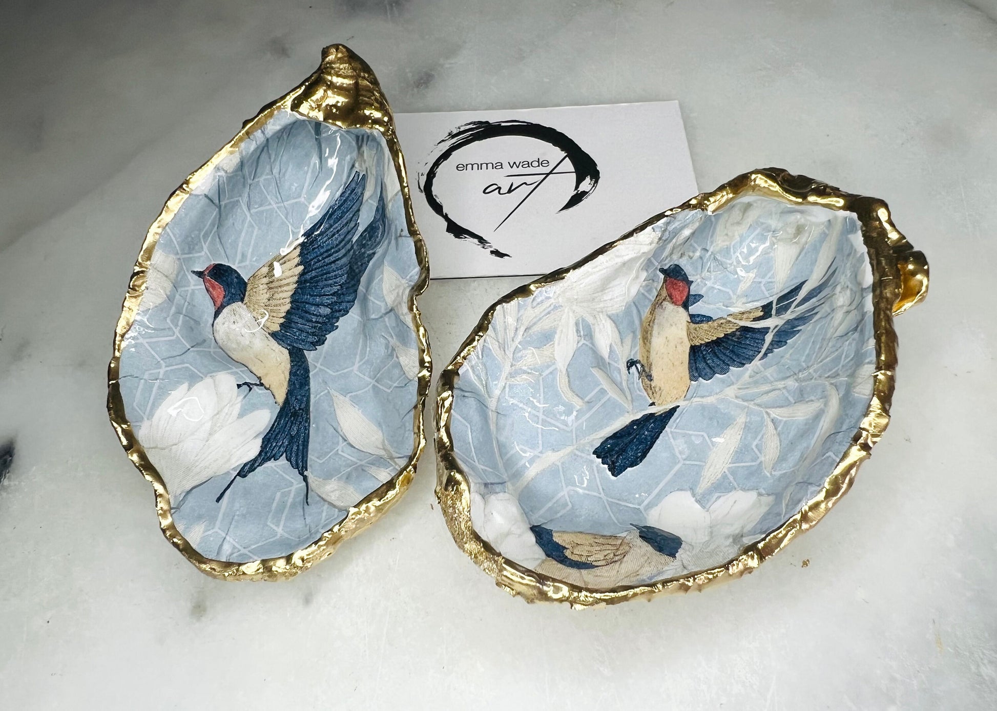 Two natural oyster shell salt and pepper dishes decorated with a blue bird decoupage design, finished with hand-gilded gold leaf edges and sealed with glossy resin. Shown with small gold spoons, these handmade coastal shell pinch pots are crafted from Dorset shells and styled as an elegant table setting accessory or unique gift.