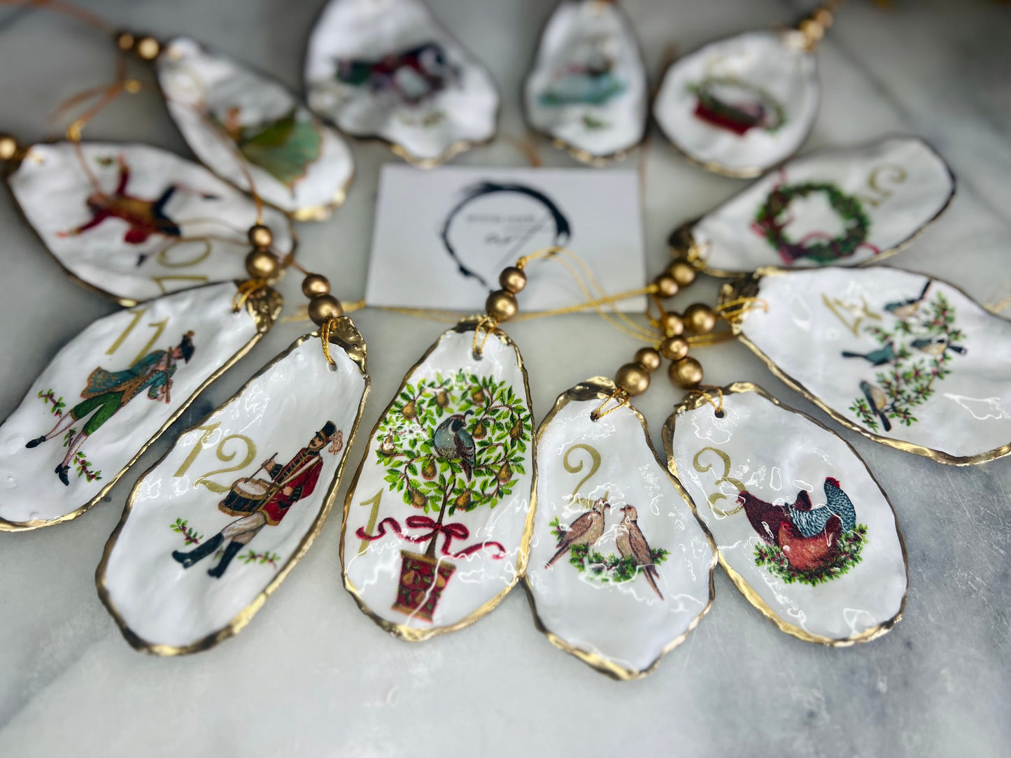 Handmade Twelve Days of Christmas oyster shell ornament set featuring all twelve traditional carol illustrations, finished with gold leaf edging, glossy resin and gold hanging cords. Coastal heirloom Christmas decorations handcrafted from Dorset oyster shells.