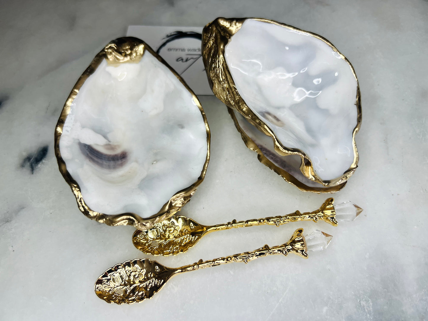 Handmade gold leaf oyster shell salt cellar set with pearl interiors and matching spoons. Coastal pinch pots made from Dorset oyster shells, sealed with glossy resin and finished with gilded edges. Luxury tableware set photographed on a light background, showing two shell salt dishes and decorative spoons, perfect for serving salt or spices at the table.