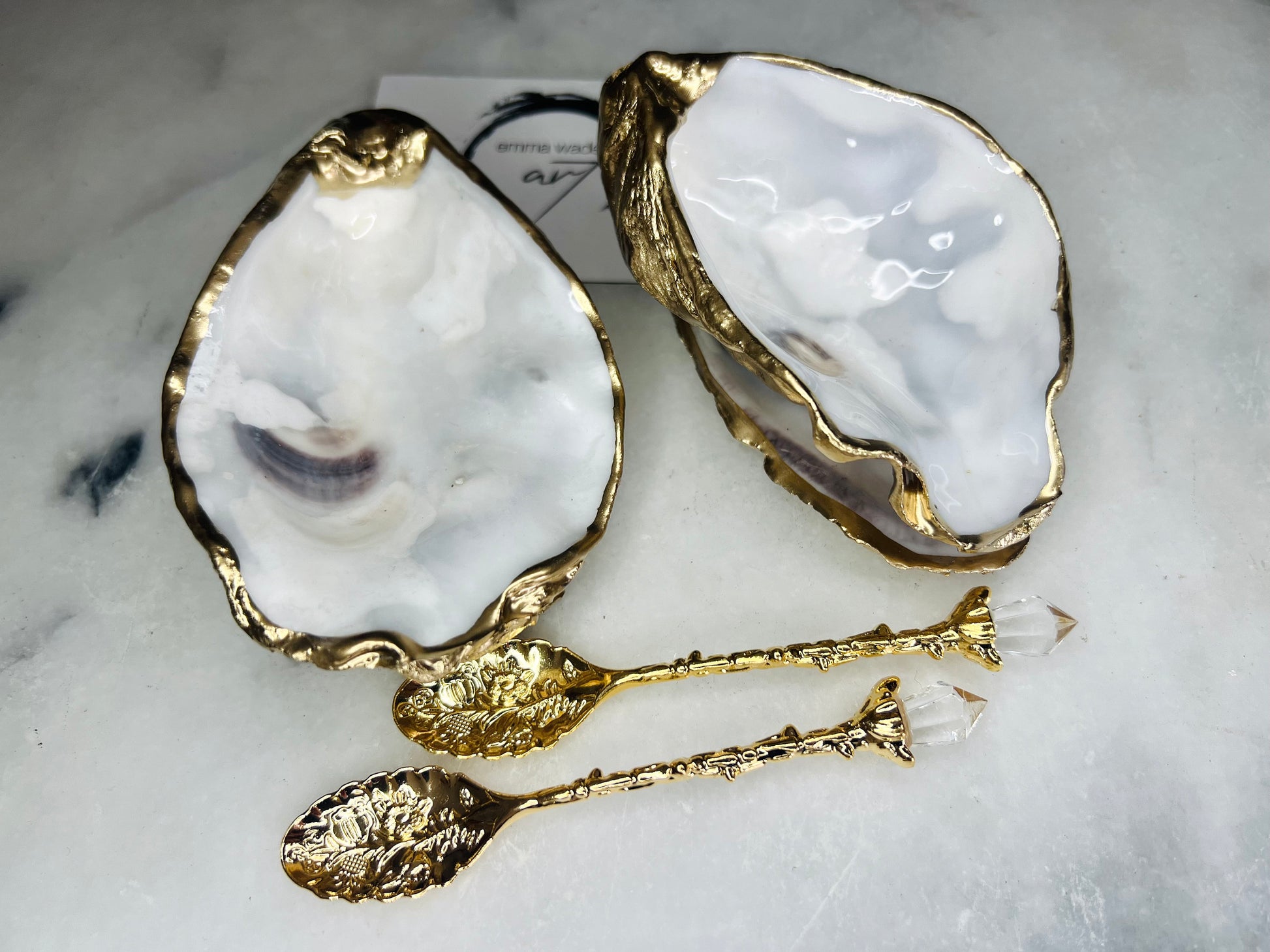 Handmade gold leaf oyster shell salt cellar set with pearl interiors and matching spoons. Coastal pinch pots made from Dorset oyster shells, sealed with glossy resin and finished with gilded edges. Luxury tableware set photographed on a light background, showing two shell salt dishes and decorative spoons, perfect for serving salt or spices at the table.