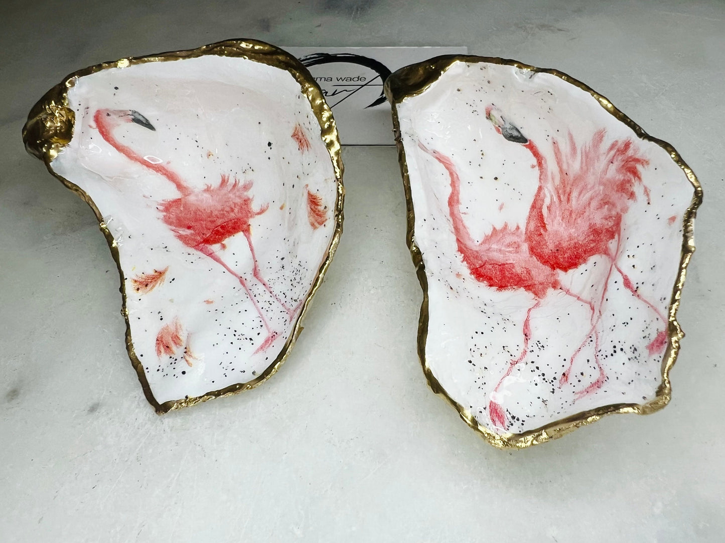 Handmade Dorset oyster shell trinket dishes featuring elegant pink flamingo bird artwork with hand-painted gold leaf edging and glossy crystal clear resin. Coastal flamingo jewellery holders styled on a white marble background with tropical blush pink tones and luxury coastal home décor styling.
