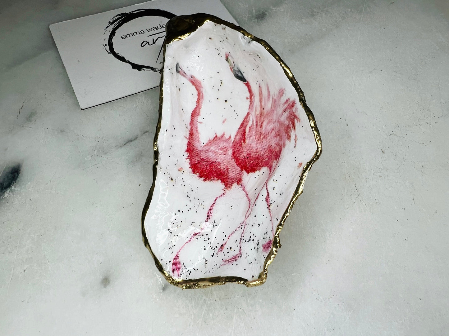 Handmade Dorset oyster shell trinket dishes featuring elegant pink flamingo bird artwork with hand-painted gold leaf edging and glossy crystal clear resin. Coastal flamingo jewellery holders styled on a white marble background with tropical blush pink tones and luxury coastal home décor styling.