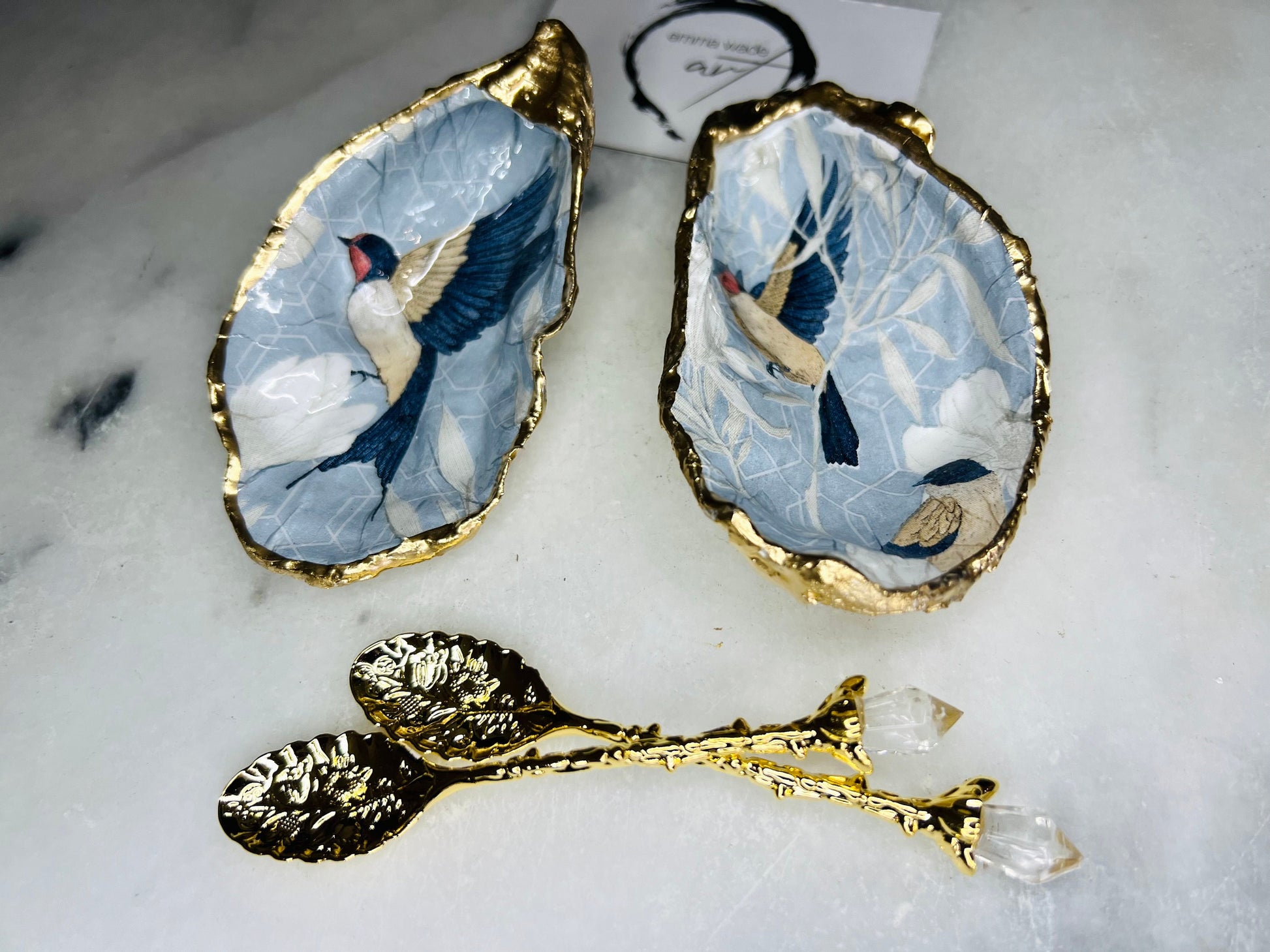 Two natural oyster shell salt and pepper dishes decorated with a blue bird decoupage design, finished with hand-gilded gold leaf edges and sealed with glossy resin. Shown with small gold spoons, these handmade coastal shell pinch pots are crafted from Dorset shells and styled as an elegant table setting accessory or unique gift.