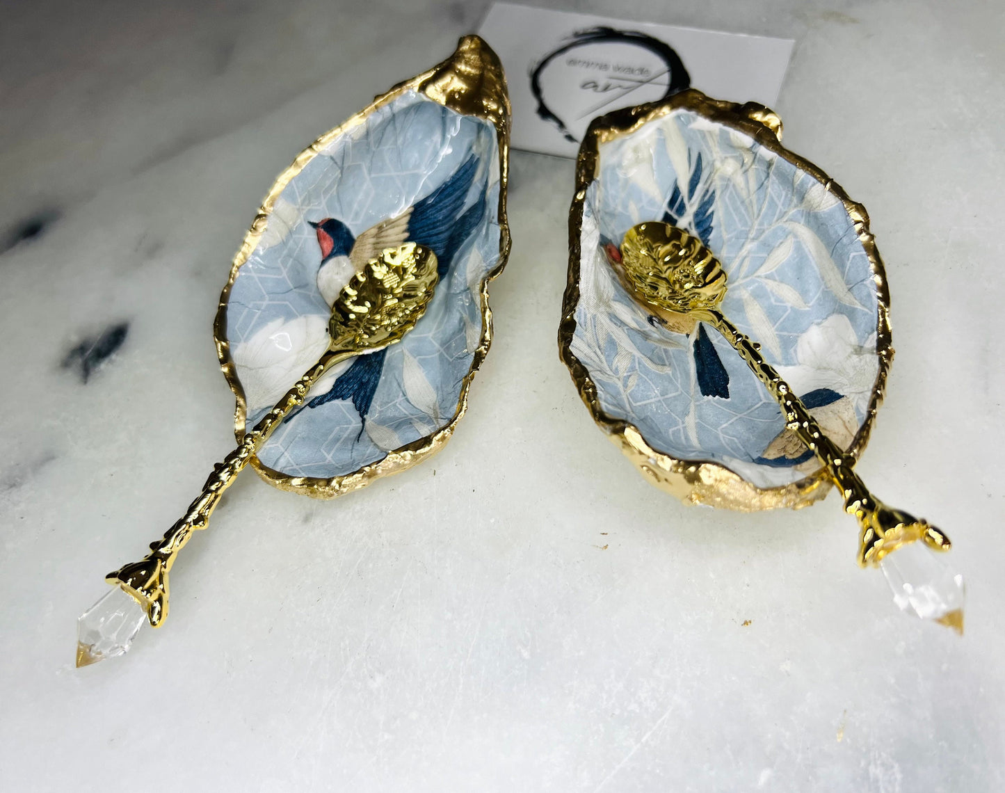 Two natural oyster shell salt and pepper dishes decorated with a blue bird decoupage design, finished with hand-gilded gold leaf edges and sealed with glossy resin. Shown with small gold spoons, these handmade coastal shell pinch pots are crafted from Dorset shells and styled as an elegant table setting accessory or unique gift.