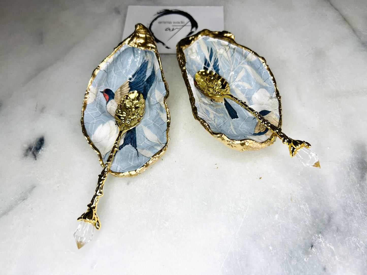 Two natural oyster shell salt and pepper dishes decorated with a blue bird decoupage design, finished with hand-gilded gold leaf edges and sealed with glossy resin. Shown with small gold spoons, these handmade coastal shell pinch pots are crafted from Dorset shells and styled as an elegant table setting accessory or unique gift.