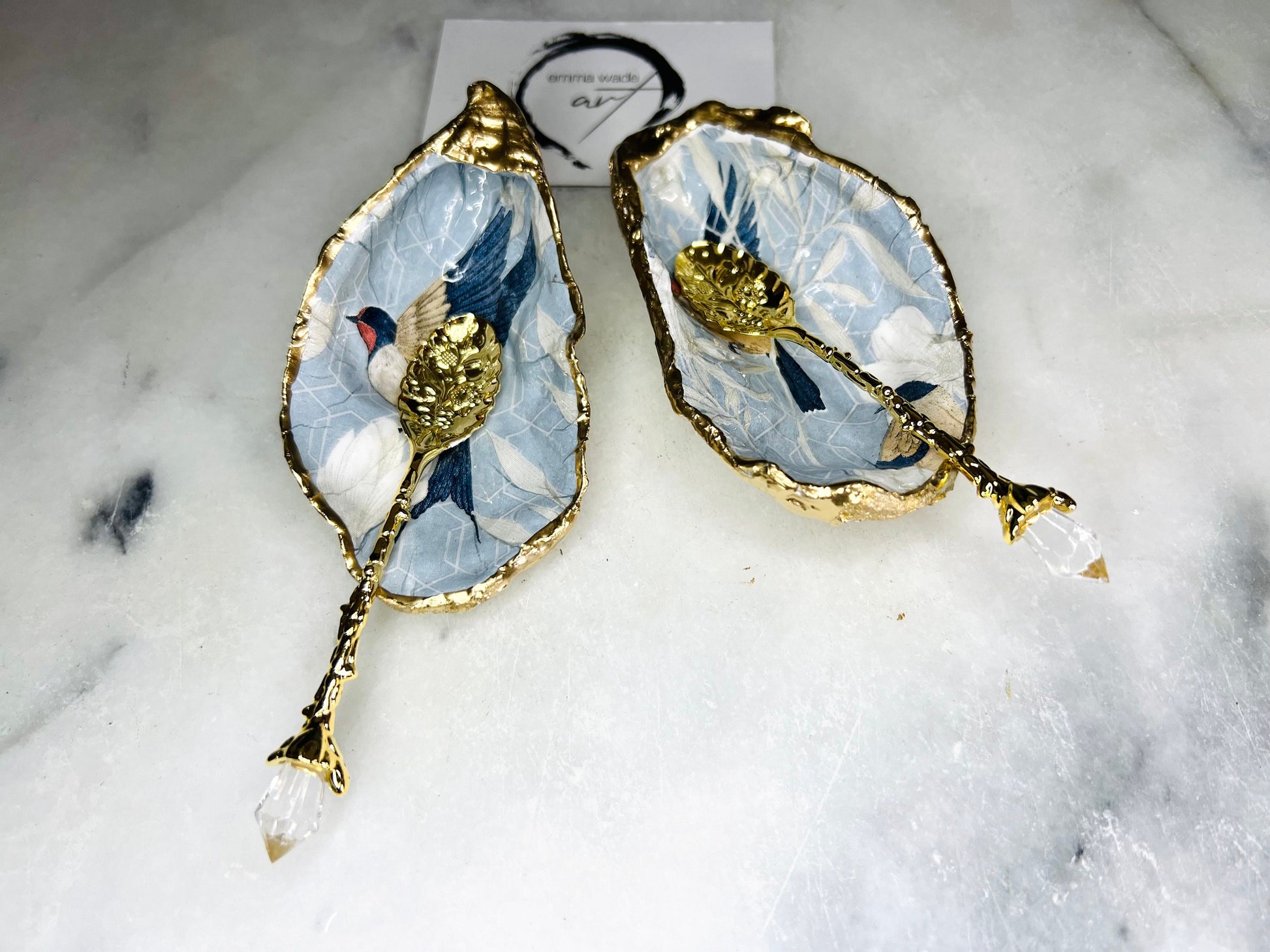 Two natural oyster shell salt and pepper dishes decorated with a blue bird decoupage design, finished with hand-gilded gold leaf edges and sealed with glossy resin. Shown with small gold spoons, these handmade coastal shell pinch pots are crafted from Dorset shells and styled as an elegant table setting accessory or unique gift.