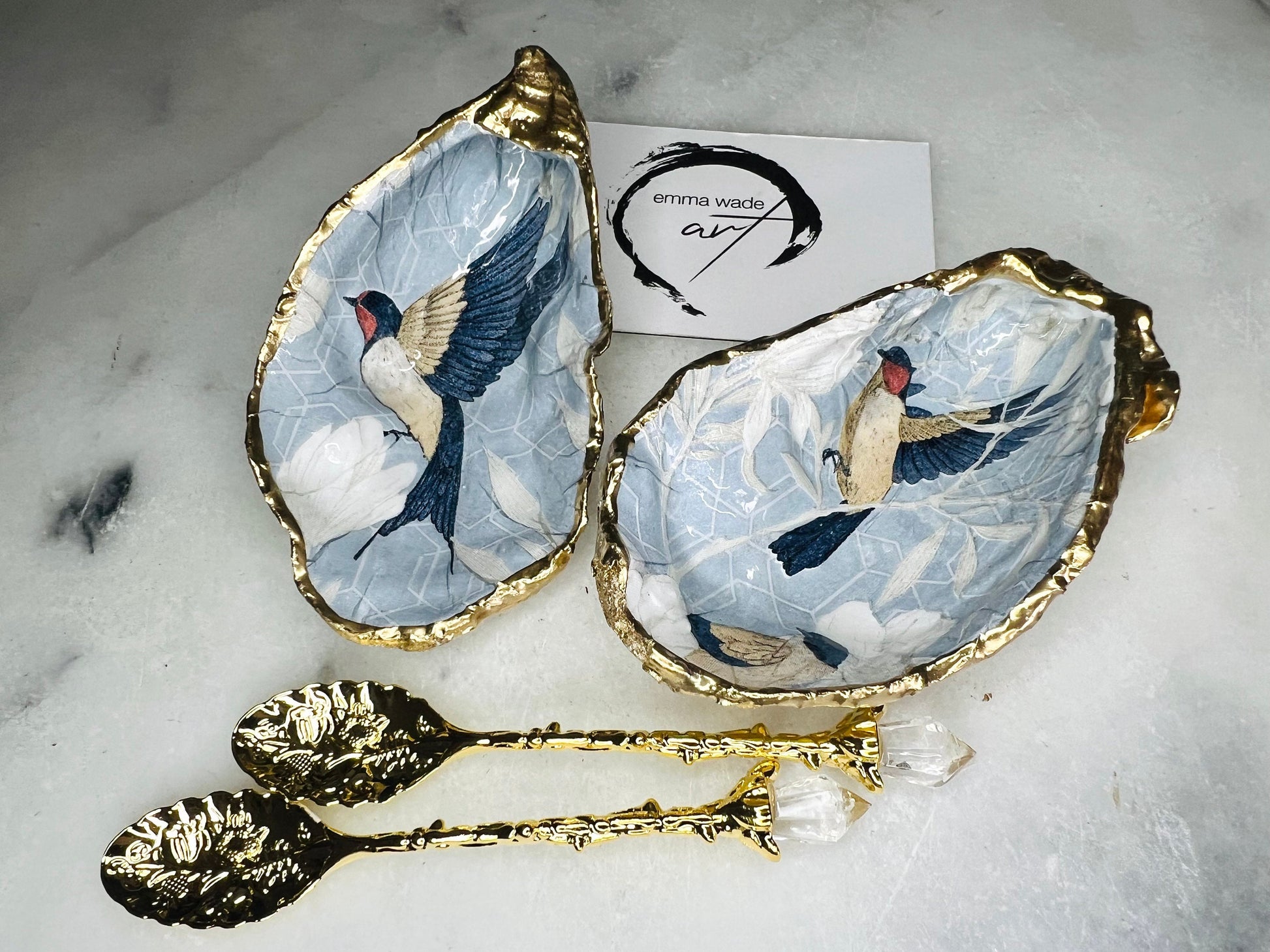 Two natural oyster shell salt and pepper dishes decorated with a blue bird decoupage design, finished with hand-gilded gold leaf edges and sealed with glossy resin. Shown with small gold spoons, these handmade coastal shell pinch pots are crafted from Dorset shells and styled as an elegant table setting accessory or unique gift.