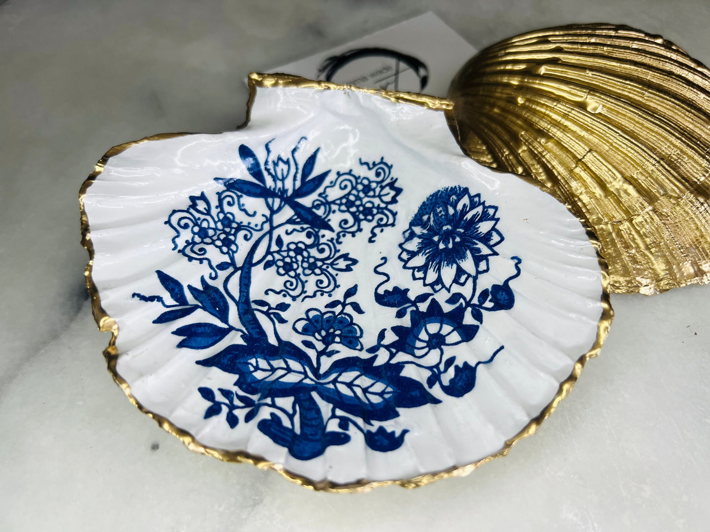 Handmade blue onion floral scallop shell trinket dish with gold leaf edging, shown on a light background. Crafted from a natural Dorset scallop shell, the dish features a classic blue and white floral pattern inspired by traditional onion designs, sealed with glossy epoxy resin. Designed as a jewellery or ring holder, this decorative shell dish is finished by hand and each piece is unique.