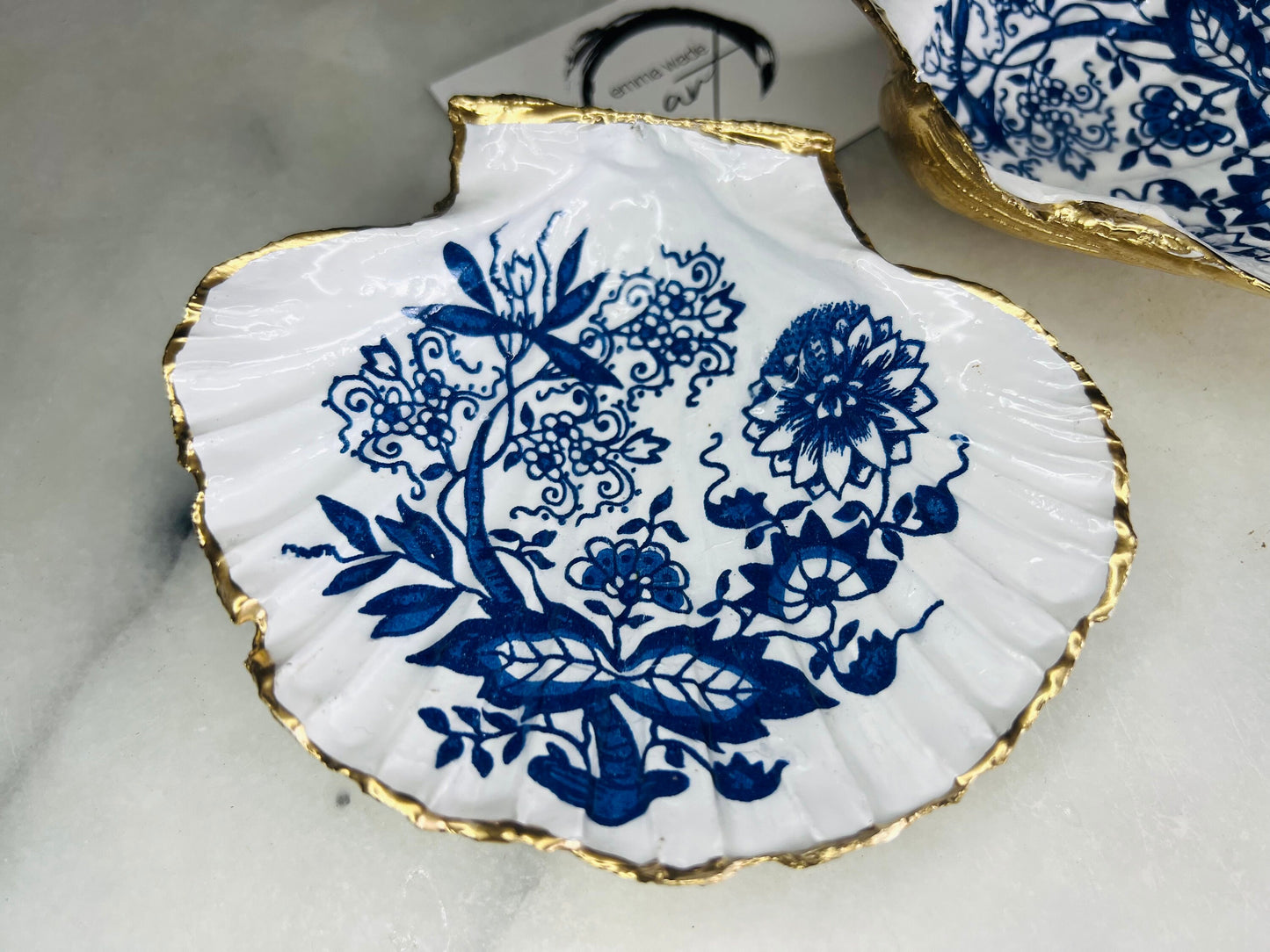 Handmade blue onion floral scallop shell trinket dish with gold leaf edging, shown on a light background. Crafted from a natural Dorset scallop shell, the dish features a classic blue and white floral pattern inspired by traditional onion designs, sealed with glossy epoxy resin. Designed as a jewellery or ring holder, this decorative shell dish is finished by hand and each piece is unique.