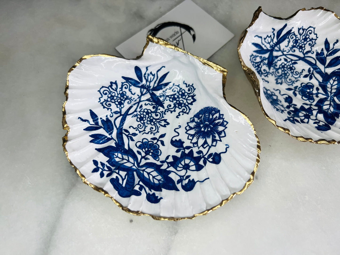 Handmade blue onion floral scallop shell trinket dish with gold leaf edging, shown on a light background. Crafted from a natural Dorset scallop shell, the dish features a classic blue and white floral pattern inspired by traditional onion designs, sealed with glossy epoxy resin. Designed as a jewellery or ring holder, this decorative shell dish is finished by hand and each piece is unique.