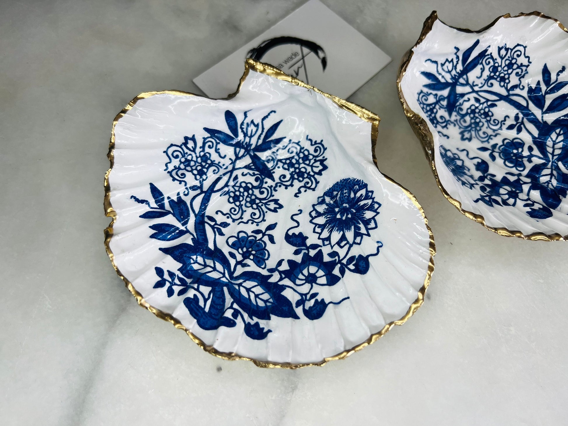 Handmade blue onion floral scallop shell trinket dish with gold leaf edging, shown on a light background. Crafted from a natural Dorset scallop shell, the dish features a classic blue and white floral pattern inspired by traditional onion designs, sealed with glossy epoxy resin. Designed as a jewellery or ring holder, this decorative shell dish is finished by hand and each piece is unique.