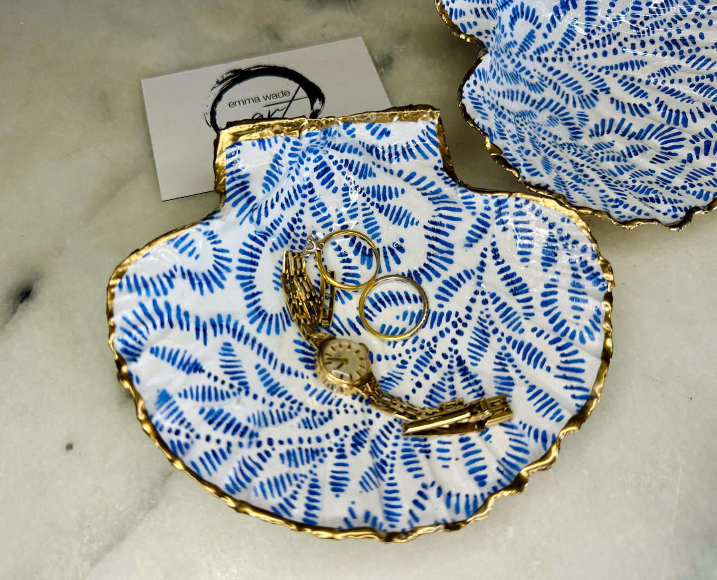 Handmade block print scallop shell trinket dish featuring a blue leaf pattern, finished with gilded gold leaf edges and sealed with a glossy resin. Crafted from a natural Dorset scallop shell, shown as a decorative jewellery tray on a light background, perfect for rings, earrings and small keepsakes. Coastal-inspired home decor, ideal for a bedside table, dressing table or bathroom, each piece one of a kind.
