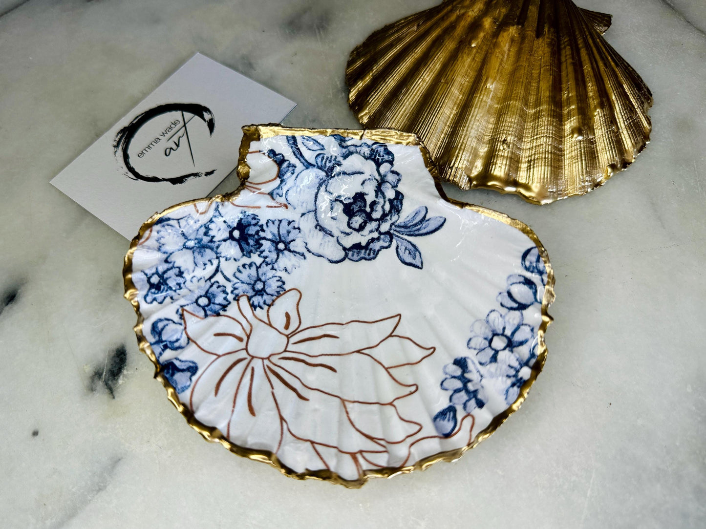 Blue and white Palmers floral scallop shell trinket dish with hand gilded liquid gold leaf edging and a glossy crystal clear resin finish. Handmade from a sustainably sourced Dorset scallop shell and designed as a luxury coastal jewellery tray, ring catcher, trinket holder or decorative shell dish. Elegant blue floral botanical pattern inspired by vintage ceramics and coastal interiors, styled for dressing tables, bathrooms, bedside tables or beach house decor. Handmade artisan shell gift.