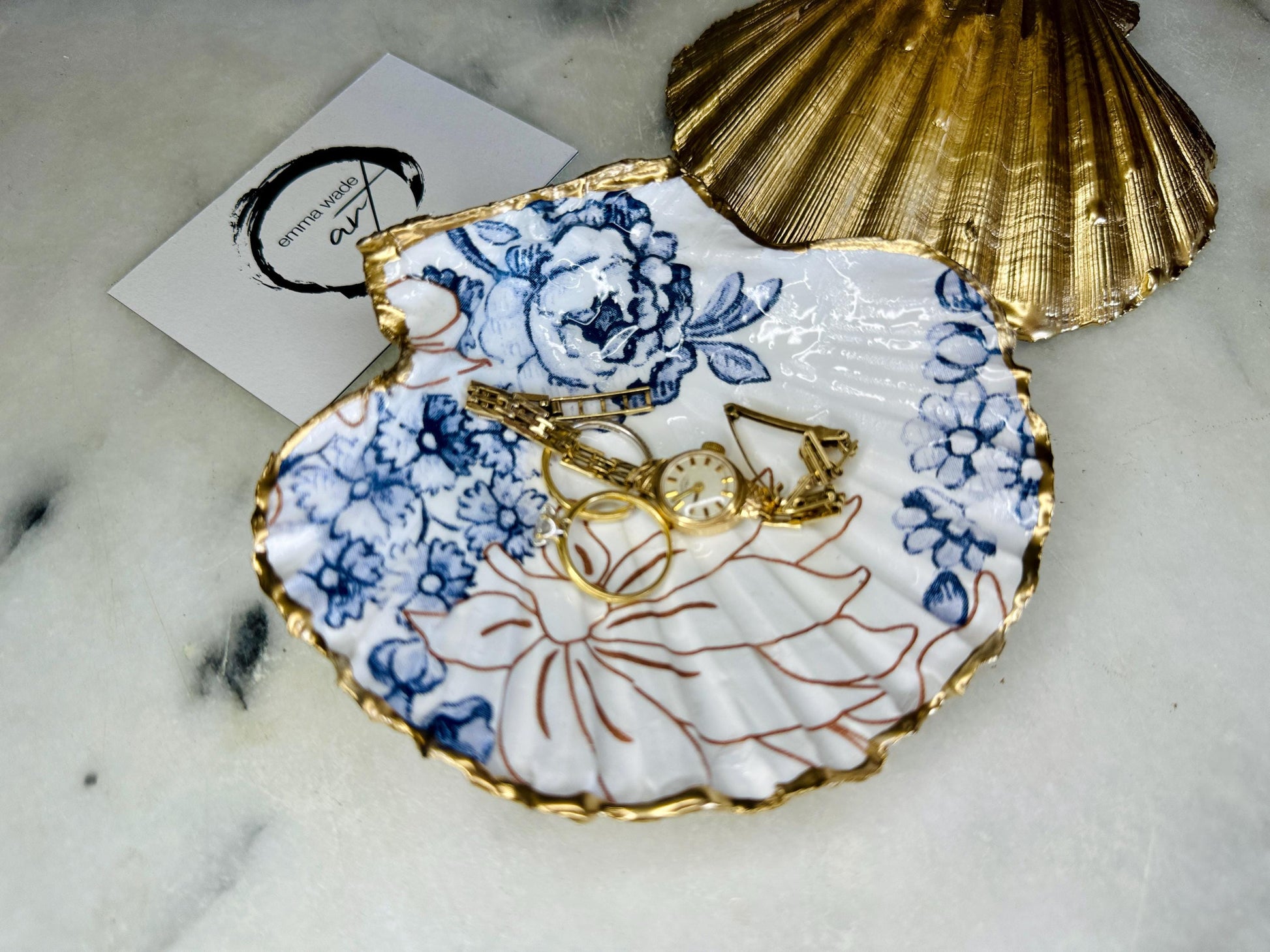 Blue and white Palmers floral scallop shell trinket dish with hand gilded liquid gold leaf edging and a glossy crystal clear resin finish. Handmade from a sustainably sourced Dorset scallop shell and designed as a luxury coastal jewellery tray, ring catcher, trinket holder or decorative shell dish. Elegant blue floral botanical pattern inspired by vintage ceramics and coastal interiors, styled for dressing tables, bathrooms, bedside tables or beach house decor. Handmade artisan shell gift.