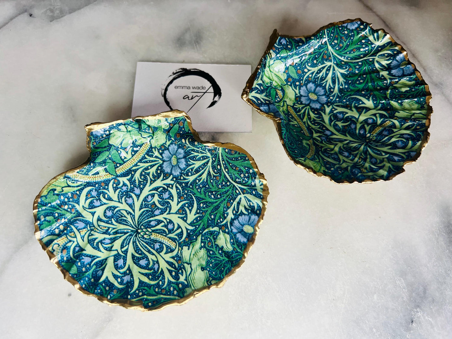 Handmade William Morris inspired scallop shell trinket dish in green and blue tones, featuring a seaweed decoupage design. Finished with gold leaf and sealed with glossy resin. Coastal jewellery dish made from a UK scallop shell, perfect for holding rings, earrings and small treasures. Decorative shell bowl photographed on a light background, showing the natural shell shape and gilded edges.