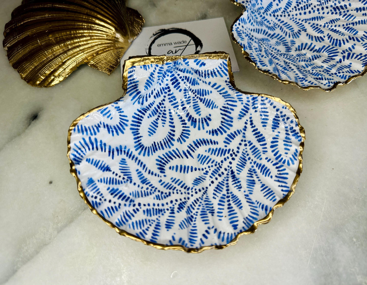 Handmade block print scallop shell trinket dish featuring a blue leaf pattern, finished with gilded gold leaf edges and sealed with a glossy resin. Crafted from a natural Dorset scallop shell, shown as a decorative jewellery tray on a light background, perfect for rings, earrings and small keepsakes. Coastal-inspired home decor, ideal for a bedside table, dressing table or bathroom, each piece one of a kind.