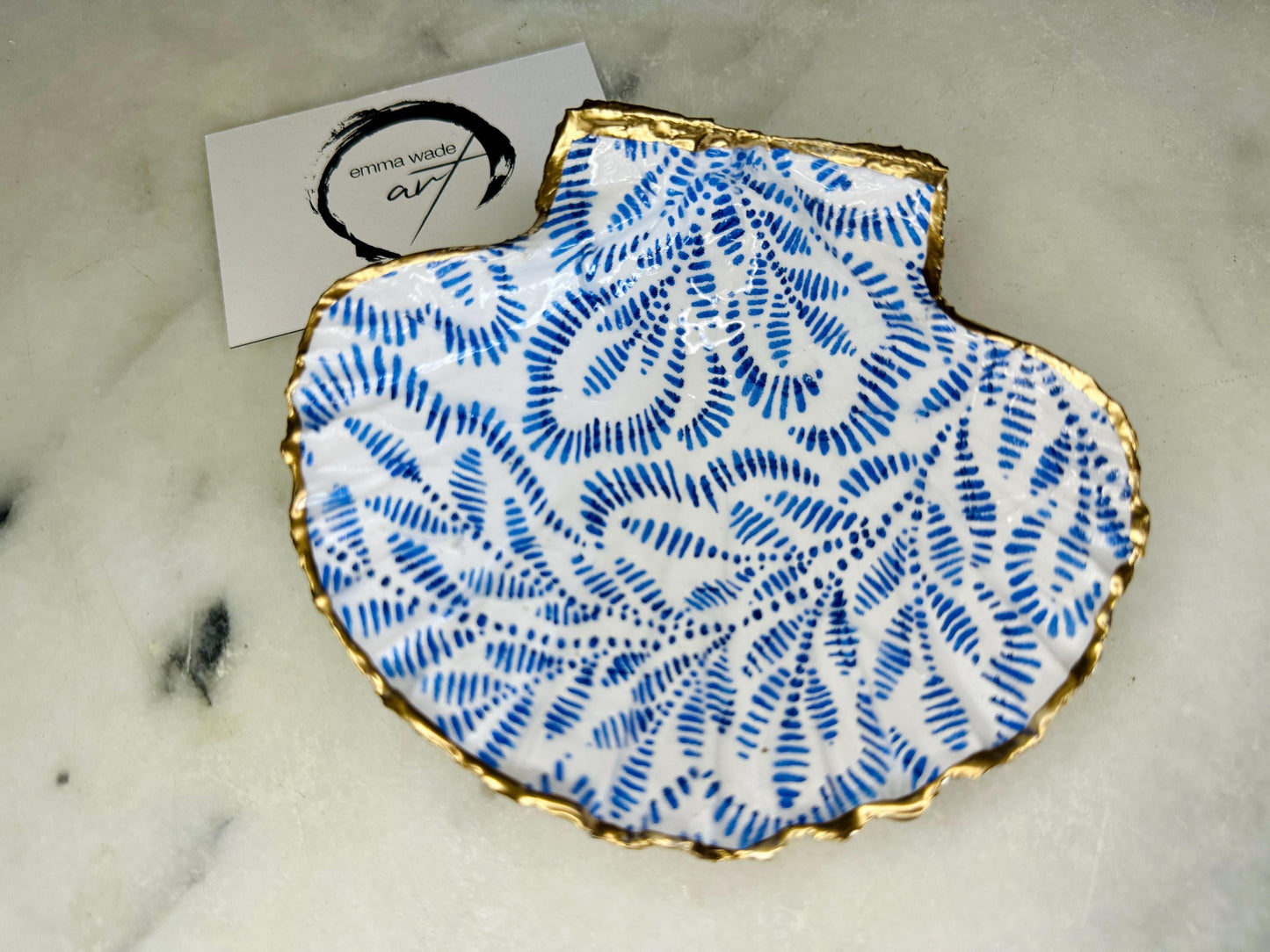 Handmade block print scallop shell trinket dish featuring a blue leaf pattern, finished with gilded gold leaf edges and sealed with a glossy resin. Crafted from a natural Dorset scallop shell, shown as a decorative jewellery tray on a light background, perfect for rings, earrings and small keepsakes. Coastal-inspired home decor, ideal for a bedside table, dressing table or bathroom, each piece one of a kind.