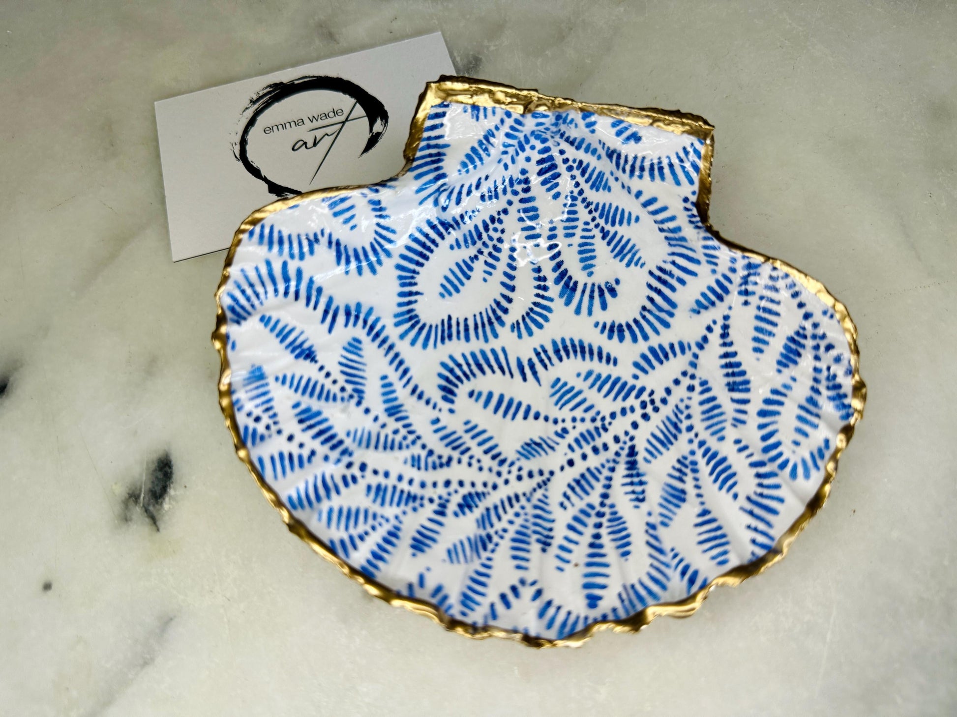 Handmade block print scallop shell trinket dish featuring a blue leaf pattern, finished with gilded gold leaf edges and sealed with a glossy resin. Crafted from a natural Dorset scallop shell, shown as a decorative jewellery tray on a light background, perfect for rings, earrings and small keepsakes. Coastal-inspired home decor, ideal for a bedside table, dressing table or bathroom, each piece one of a kind.