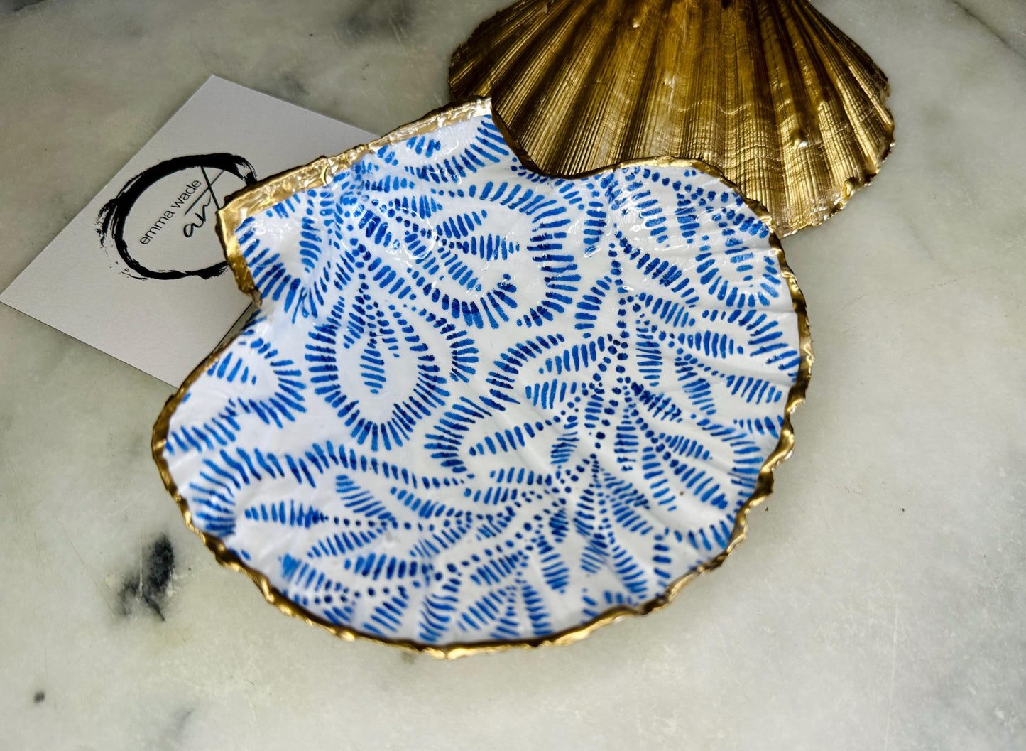 Handmade block print scallop shell trinket dish featuring a blue leaf pattern, finished with gilded gold leaf edges and sealed with a glossy resin. Crafted from a natural Dorset scallop shell, shown as a decorative jewellery tray on a light background, perfect for rings, earrings and small keepsakes. Coastal-inspired home decor, ideal for a bedside table, dressing table or bathroom, each piece one of a kind.