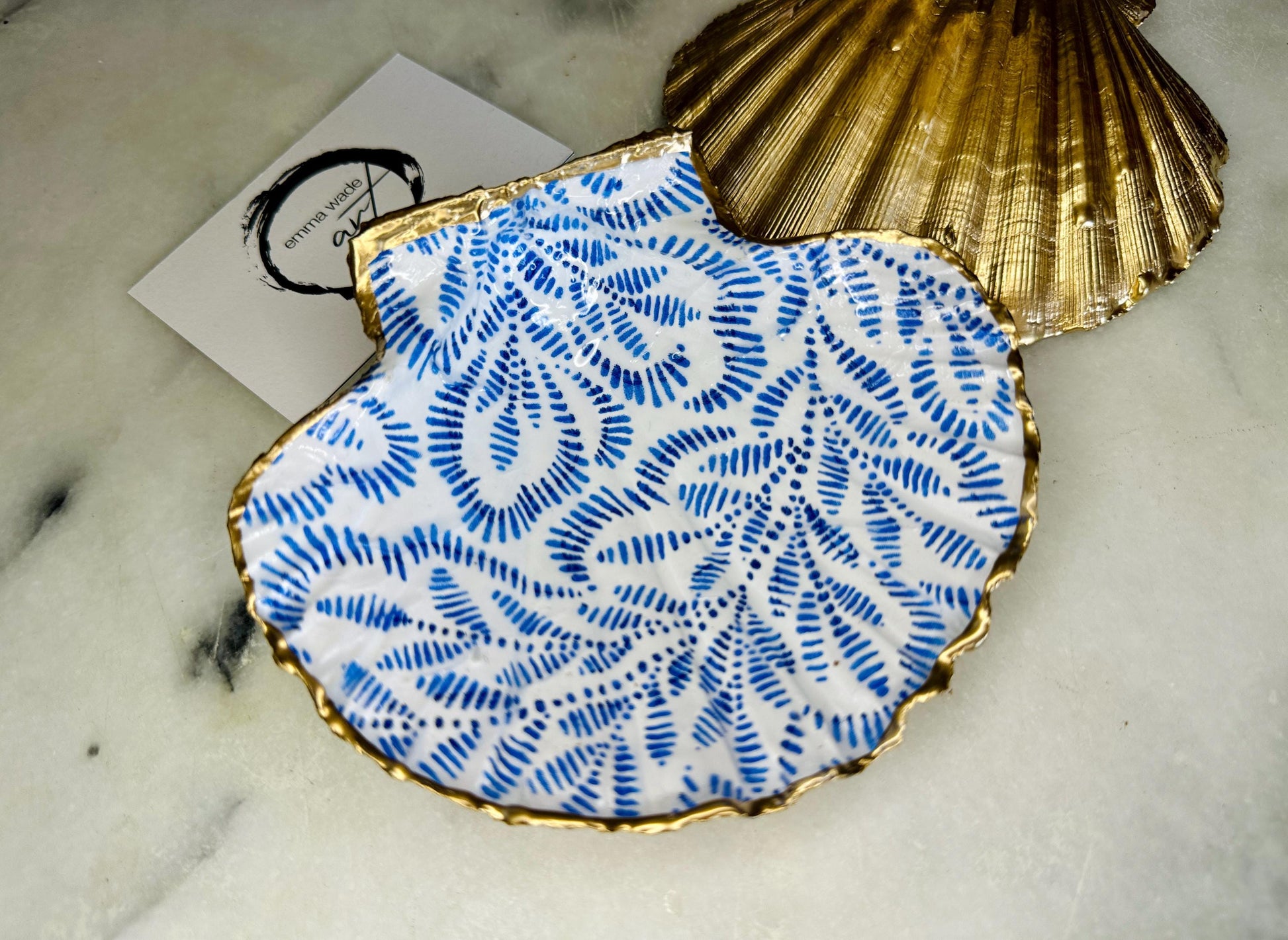 Handmade block print scallop shell trinket dish featuring a blue leaf pattern, finished with gilded gold leaf edges and sealed with a glossy resin. Crafted from a natural Dorset scallop shell, shown as a decorative jewellery tray on a light background, perfect for rings, earrings and small keepsakes. Coastal-inspired home decor, ideal for a bedside table, dressing table or bathroom, each piece one of a kind.