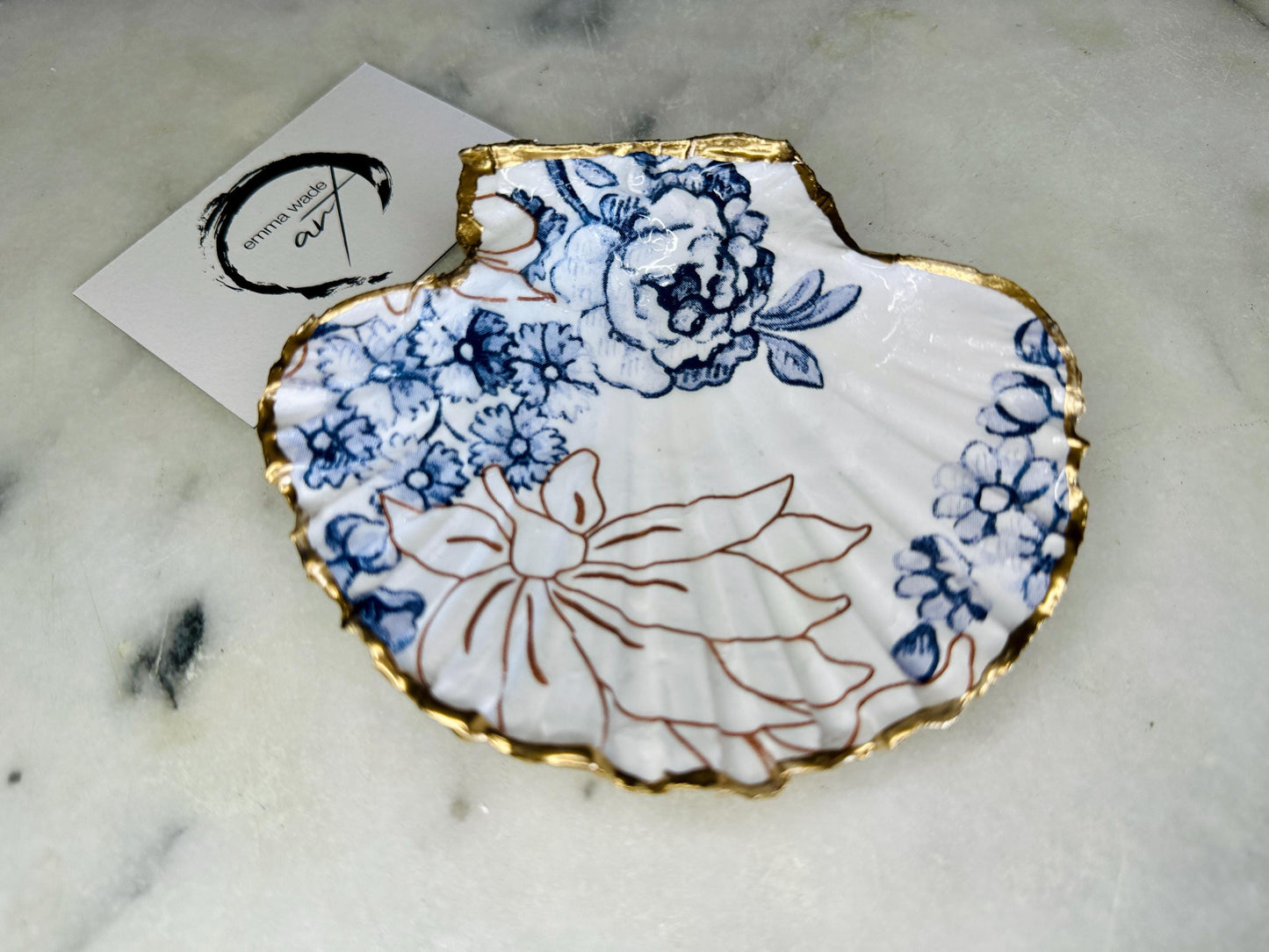 Blue and white Palmers floral scallop shell trinket dish with hand gilded liquid gold leaf edging and a glossy crystal clear resin finish. Handmade from a sustainably sourced Dorset scallop shell and designed as a luxury coastal jewellery tray, ring catcher, trinket holder or decorative shell dish. Elegant blue floral botanical pattern inspired by vintage ceramics and coastal interiors, styled for dressing tables, bathrooms, bedside tables or beach house decor. Handmade artisan shell gift.
