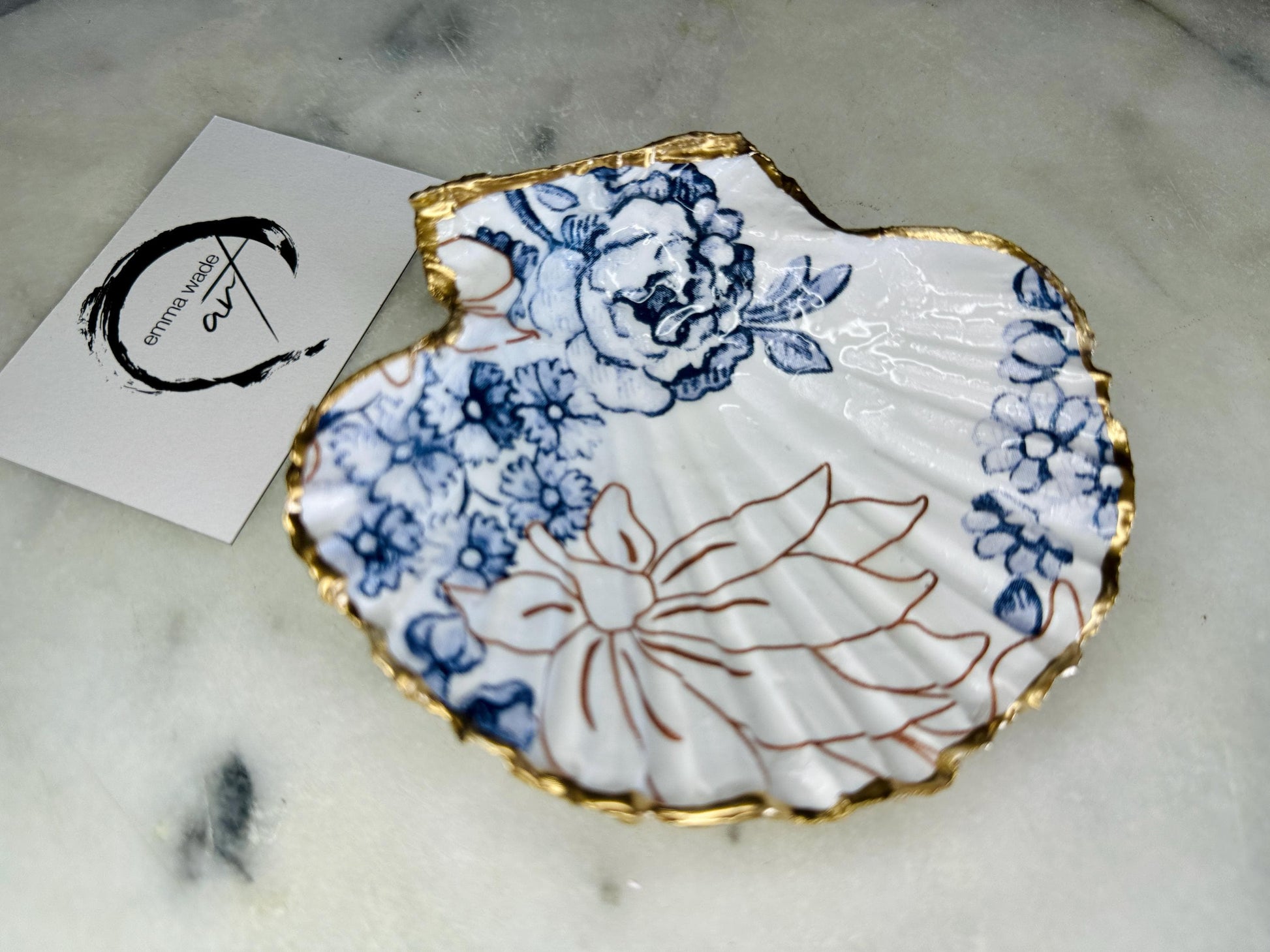 Blue and white Palmers floral scallop shell trinket dish with hand gilded liquid gold leaf edging and a glossy crystal clear resin finish. Handmade from a sustainably sourced Dorset scallop shell and designed as a luxury coastal jewellery tray, ring catcher, trinket holder or decorative shell dish. Elegant blue floral botanical pattern inspired by vintage ceramics and coastal interiors, styled for dressing tables, bathrooms, bedside tables or beach house decor. Handmade artisan shell gift.