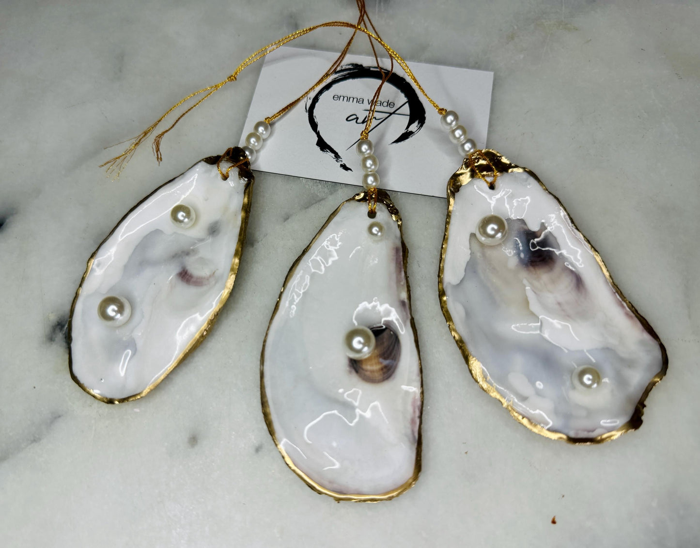 Luxury flat pearl oyster shell hanging ornaments handcrafted from natural Dorset oyster shells with glossy pearlescent interiors, two Swarovski pearls per ornament and hand-painted gold leaf edging. Elegant coastal hanging decor finished in crystal clear resin with gold hanging cord and pearl bead details. Available individually or as a set of three. Perfect for beach house interiors, luxury bathroom styling, wedding gifts, coastal Christmas decor and year-round shell home decor.