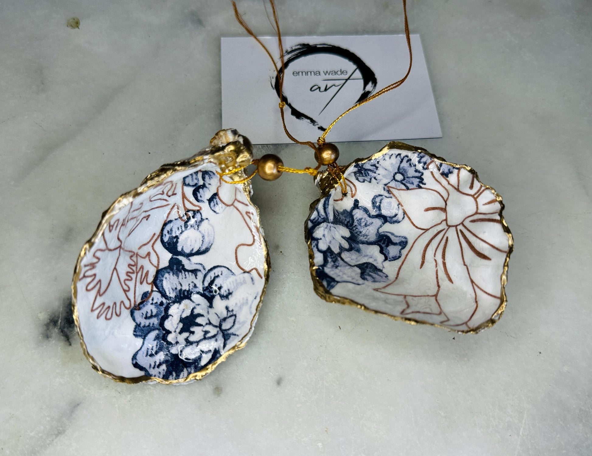 Handmade blue floral oyster shell hanging ornament shown as a single piece, decoupaged with a Palmer’s print style floral design, hand-gilded with gold leaf and sealed with glossy resin. Crafted from a natural Dorset oyster shell and finished with a gold hanging string. Coastal-inspired decorative ornament, perfect for Christmas trees, botanical home decor or a unique handmade gift. Each shell is one of a kind.