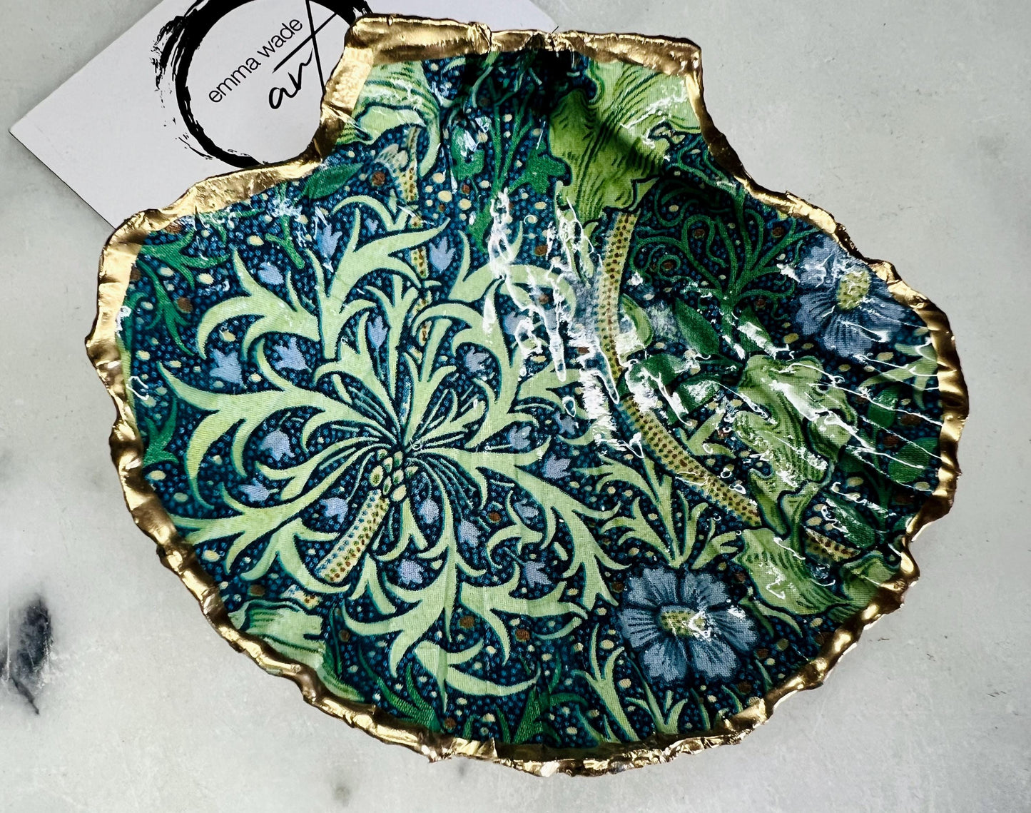Handmade William Morris inspired scallop shell trinket dish in green and blue tones, featuring a seaweed decoupage design. Finished with gold leaf and sealed with glossy resin. Coastal jewellery dish made from a UK scallop shell, perfect for holding rings, earrings and small treasures. Decorative shell bowl photographed on a light background, showing the natural shell shape and gilded edges.