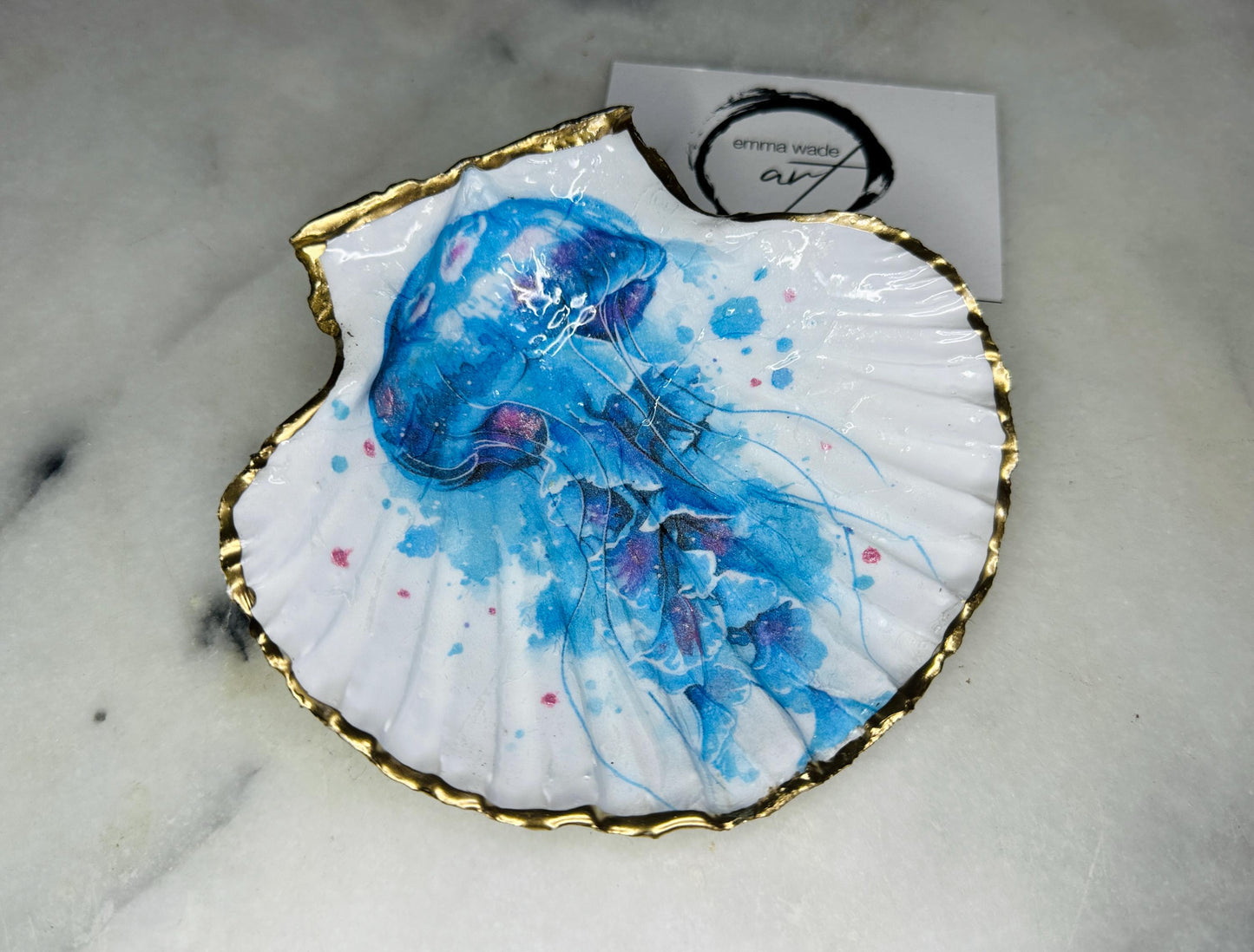 Hand illustrated blue jellyfish scallop shell trinket dish featuring original Emma Wade ART watercolour artwork. Natural Dorset scallop shell finished with hand-gilded gold leaf edging and crystal-clear resin gloss. Coastal jewellery dish, ring holder, trinket tray and ocean-inspired home decor gift. Blue and pink jellyfish design with luxury gold finish, handmade in Surrey, UK.