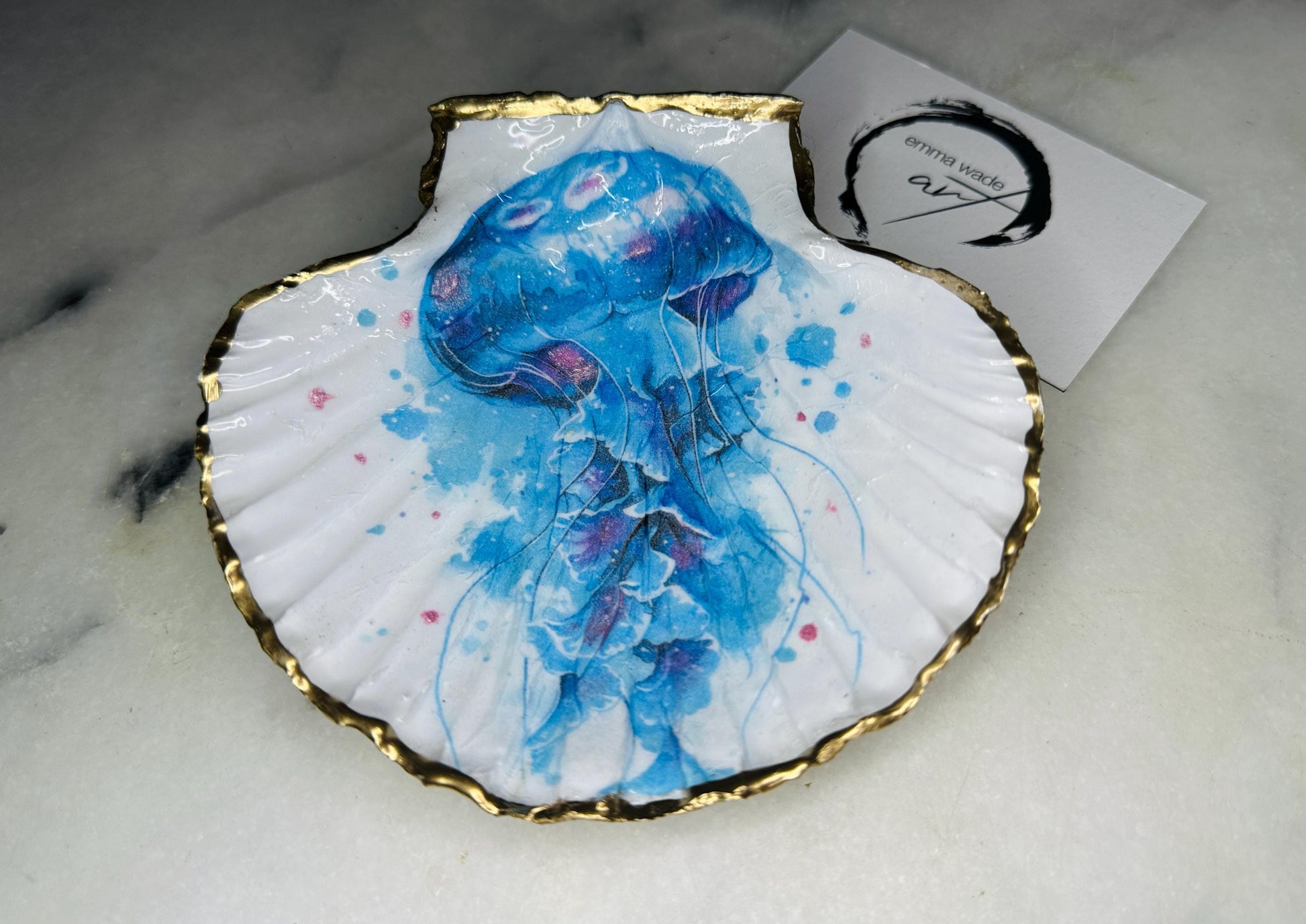 Hand illustrated blue jellyfish scallop shell trinket dish featuring original Emma Wade ART watercolour artwork. Natural Dorset scallop shell finished with hand-gilded gold leaf edging and crystal-clear resin gloss. Coastal jewellery dish, ring holder, trinket tray and ocean-inspired home decor gift. Blue and pink jellyfish design with luxury gold finish, handmade in Surrey, UK.