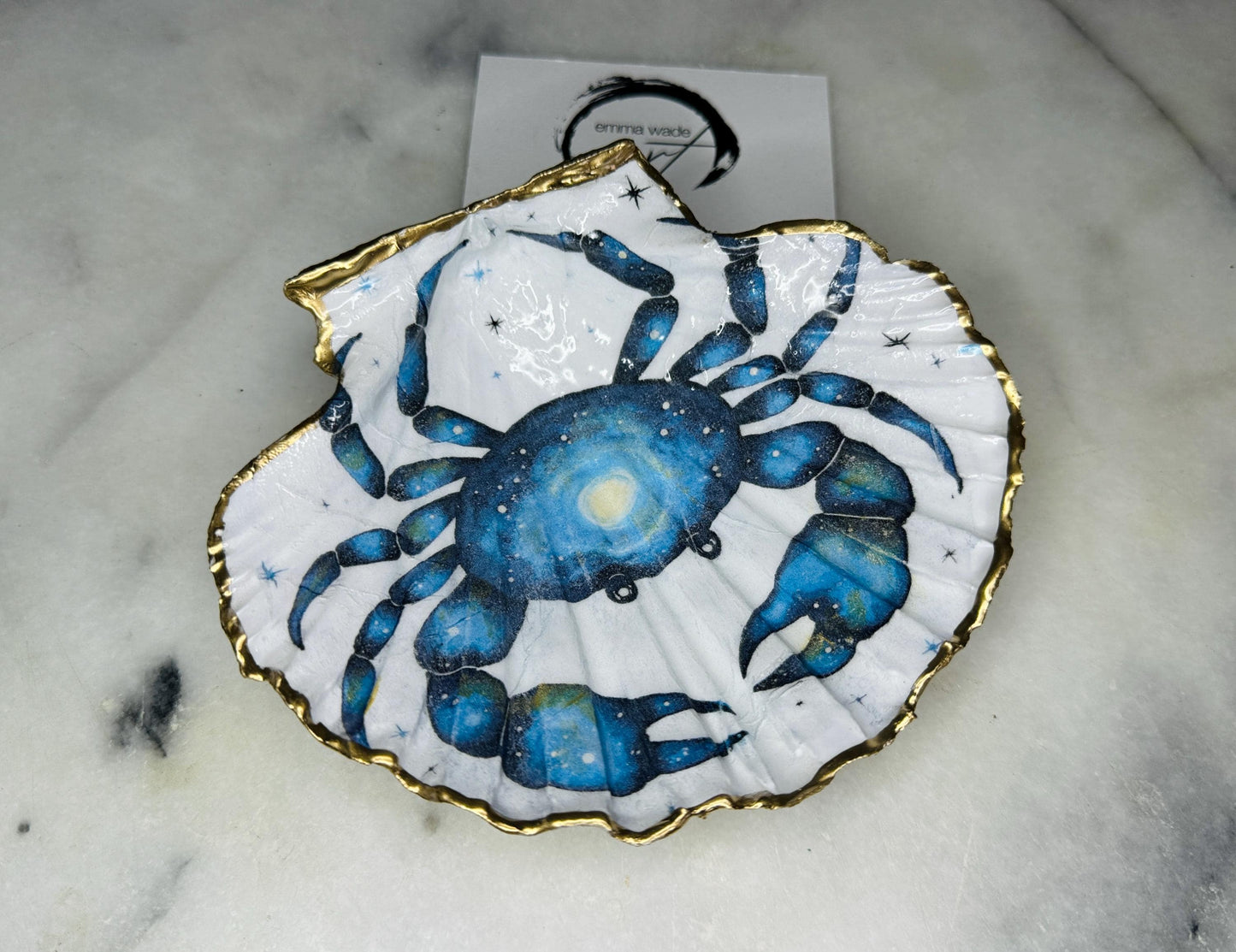 Scallop shell trinket dish with a hand drawn and hand painted blue crab design from original watercolour artwork. The artwork is applied as a decoupage print, then enhanced with painted detailing for a unique finish. Each shell is edged with liquid gold leaf and sealed with glossy resin. A handmade shell ring dish, jewellery organiser, or trinket tray for rings, earrings, or cufflinks. Perfect coastal decor or gift for weddings, anniversaries, birthdays, or Christmas.