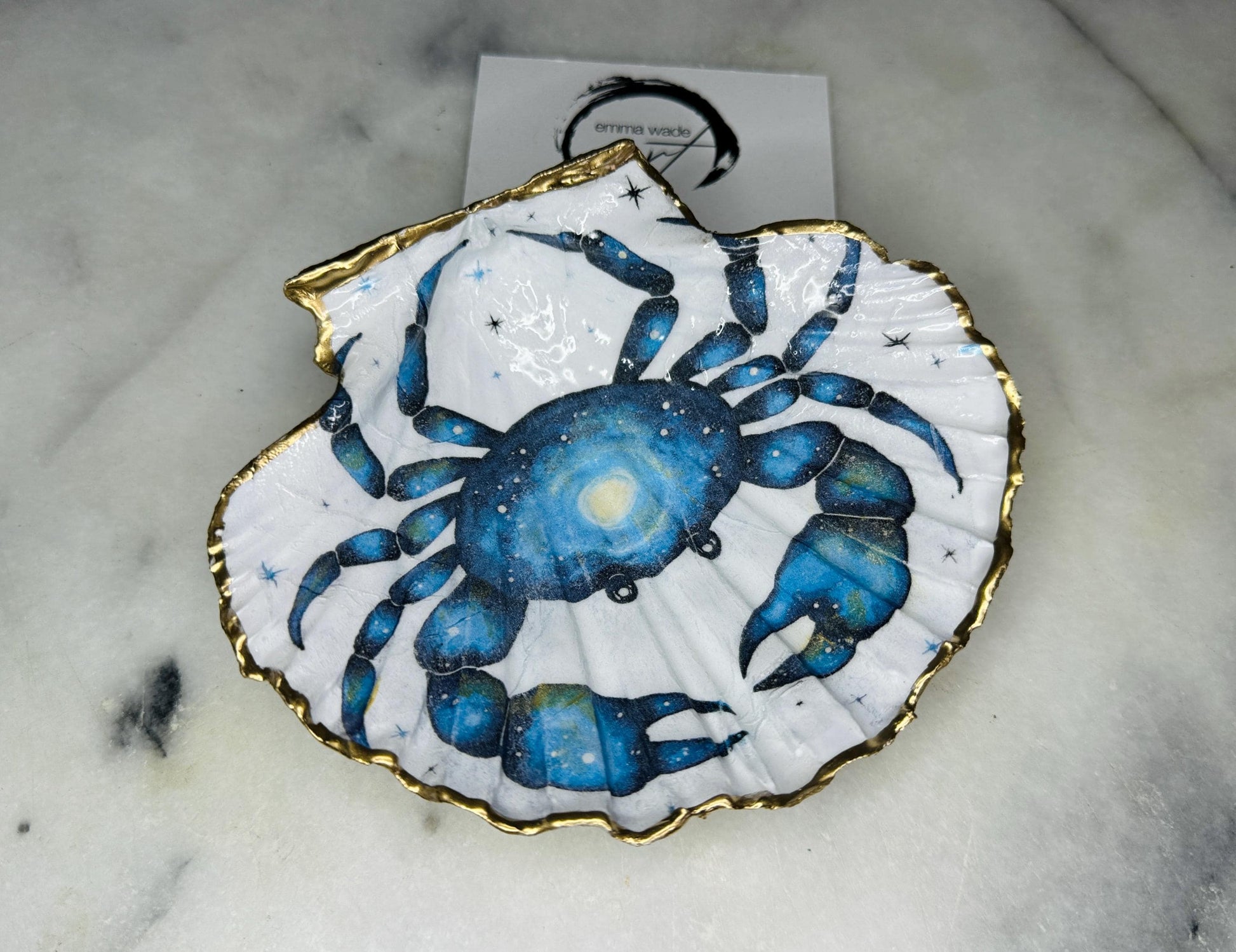 Scallop shell trinket dish with a hand drawn and hand painted blue crab design from original watercolour artwork. The artwork is applied as a decoupage print, then enhanced with painted detailing for a unique finish. Each shell is edged with liquid gold leaf and sealed with glossy resin. A handmade shell ring dish, jewellery organiser, or trinket tray for rings, earrings, or cufflinks. Perfect coastal decor or gift for weddings, anniversaries, birthdays, or Christmas.
