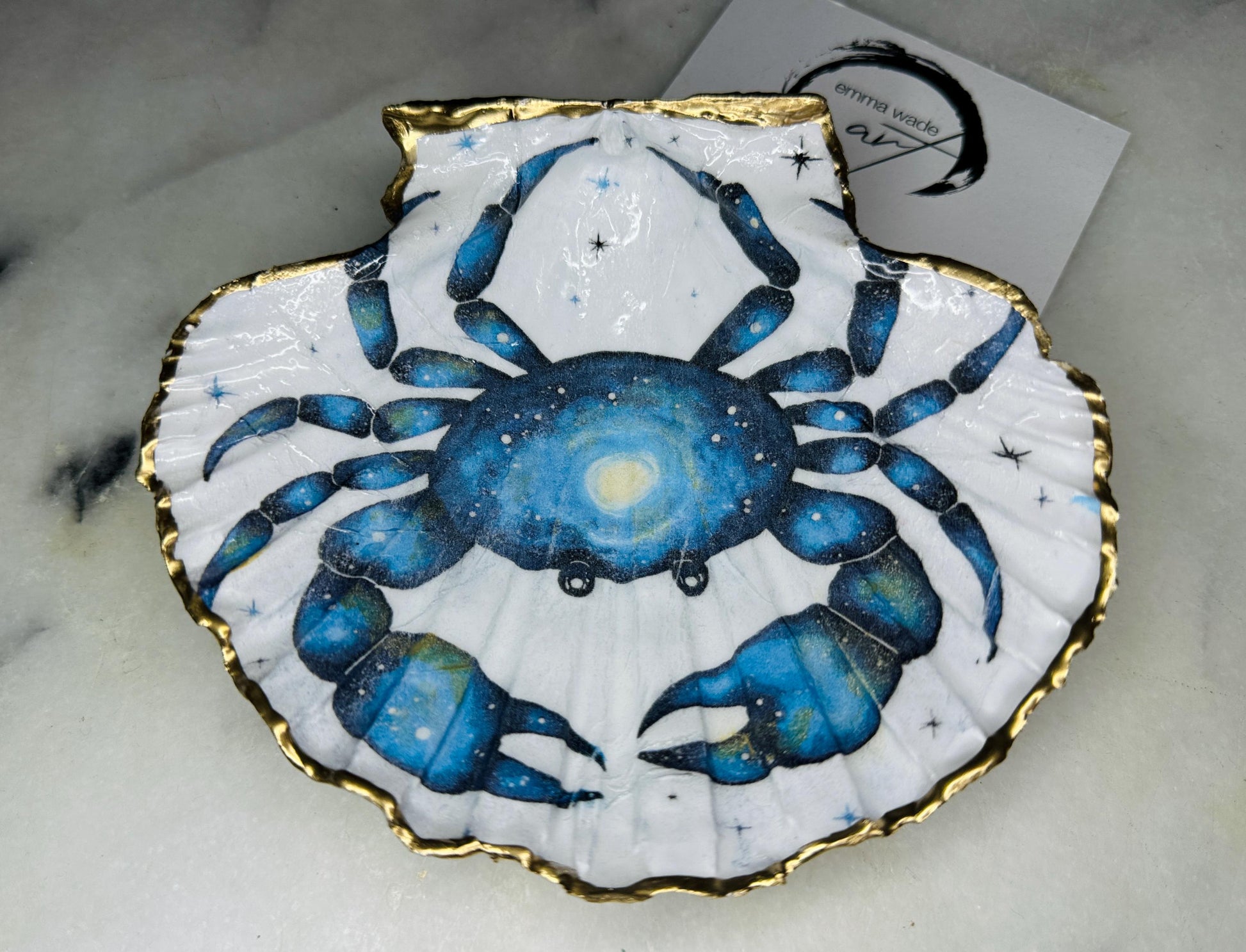 Scallop shell trinket dish with a hand drawn and hand painted blue crab design from original watercolour artwork. The artwork is applied as a decoupage print, then enhanced with painted detailing for a unique finish. Each shell is edged with liquid gold leaf and sealed with glossy resin. A handmade shell ring dish, jewellery organiser, or trinket tray for rings, earrings, or cufflinks. Perfect coastal decor or gift for weddings, anniversaries, birthdays, or Christmas.