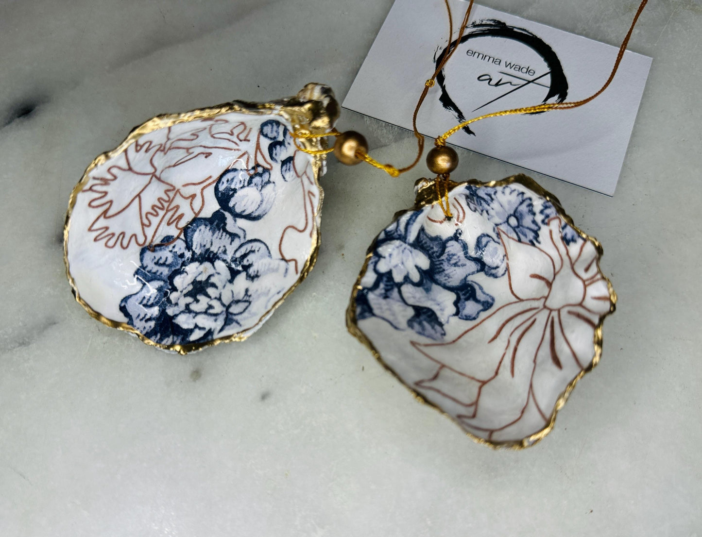 Handmade blue floral oyster shell hanging ornament shown as a single piece, decoupaged with a Palmer’s print style floral design, hand-gilded with gold leaf and sealed with glossy resin. Crafted from a natural Dorset oyster shell and finished with a gold hanging string. Coastal-inspired decorative ornament, perfect for Christmas trees, botanical home decor or a unique handmade gift. Each shell is one of a kind.