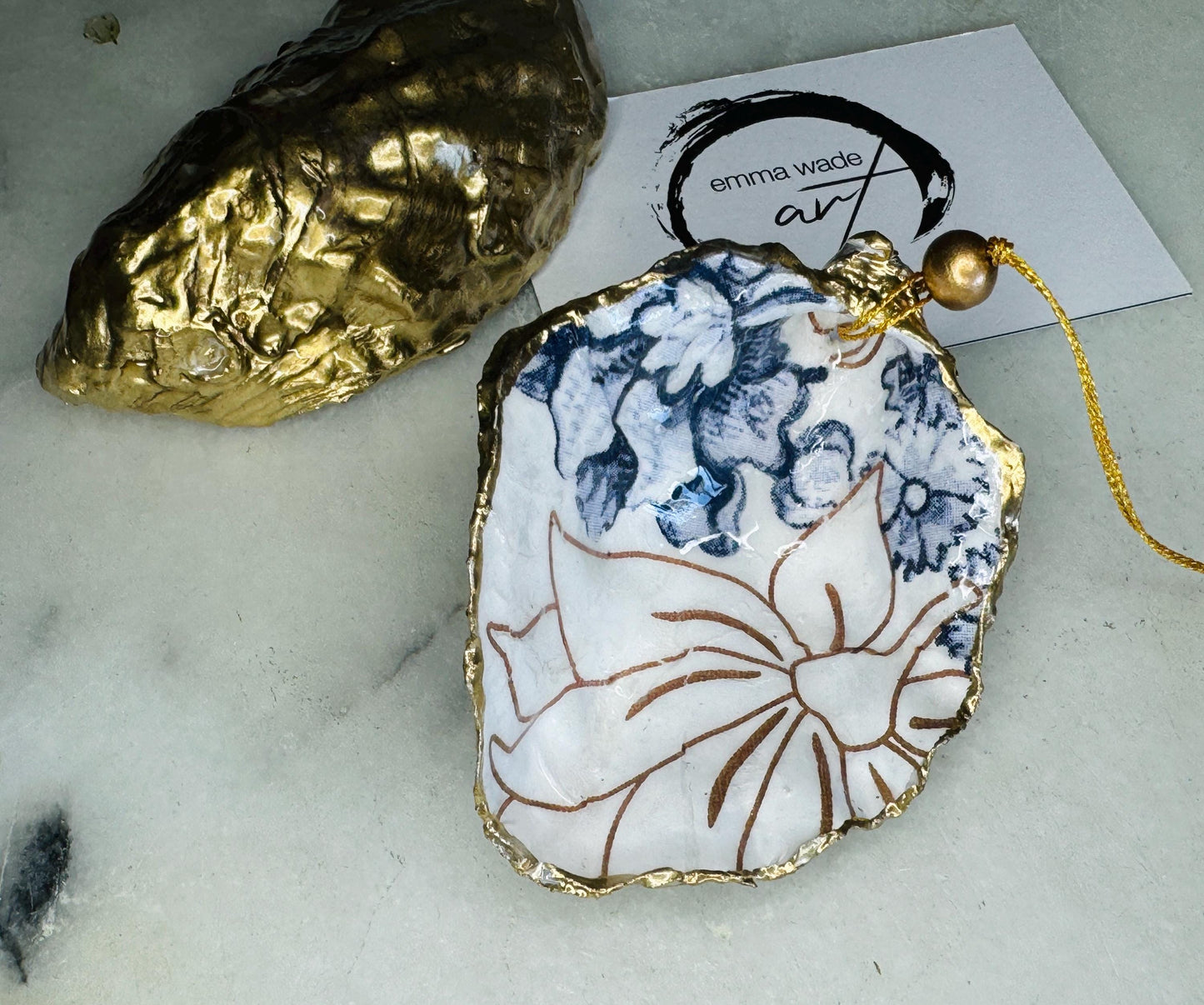 Handmade blue floral oyster shell hanging ornament shown as a single piece, decoupaged with a Palmer’s print style floral design, hand-gilded with gold leaf and sealed with glossy resin. Crafted from a natural Dorset oyster shell and finished with a gold hanging string. Coastal-inspired decorative ornament, perfect for Christmas trees, botanical home decor or a unique handmade gift. Each shell is one of a kind.
