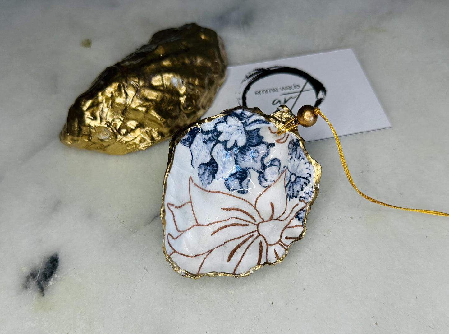 Handmade blue floral oyster shell hanging ornament shown as a single piece, decoupaged with a Palmer’s print style floral design, hand-gilded with gold leaf and sealed with glossy resin. Crafted from a natural Dorset oyster shell and finished with a gold hanging string. Coastal-inspired decorative ornament, perfect for Christmas trees, botanical home decor or a unique handmade gift. Each shell is one of a kind.