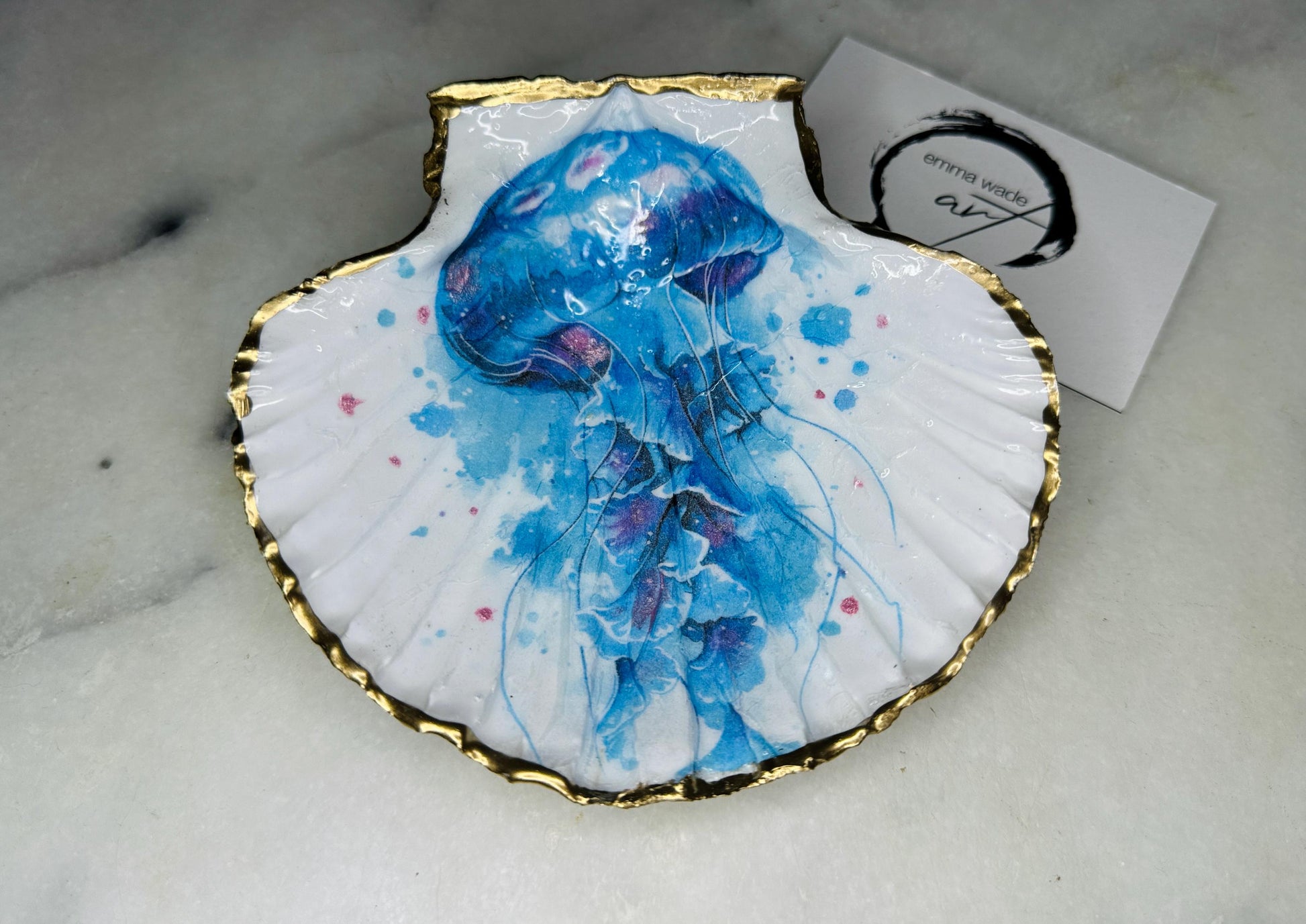 Hand illustrated blue jellyfish scallop shell trinket dish featuring original Emma Wade ART watercolour artwork. Natural Dorset scallop shell finished with hand-gilded gold leaf edging and crystal-clear resin gloss. Coastal jewellery dish, ring holder, trinket tray and ocean-inspired home decor gift. Blue and pink jellyfish design with luxury gold finish, handmade in Surrey, UK.